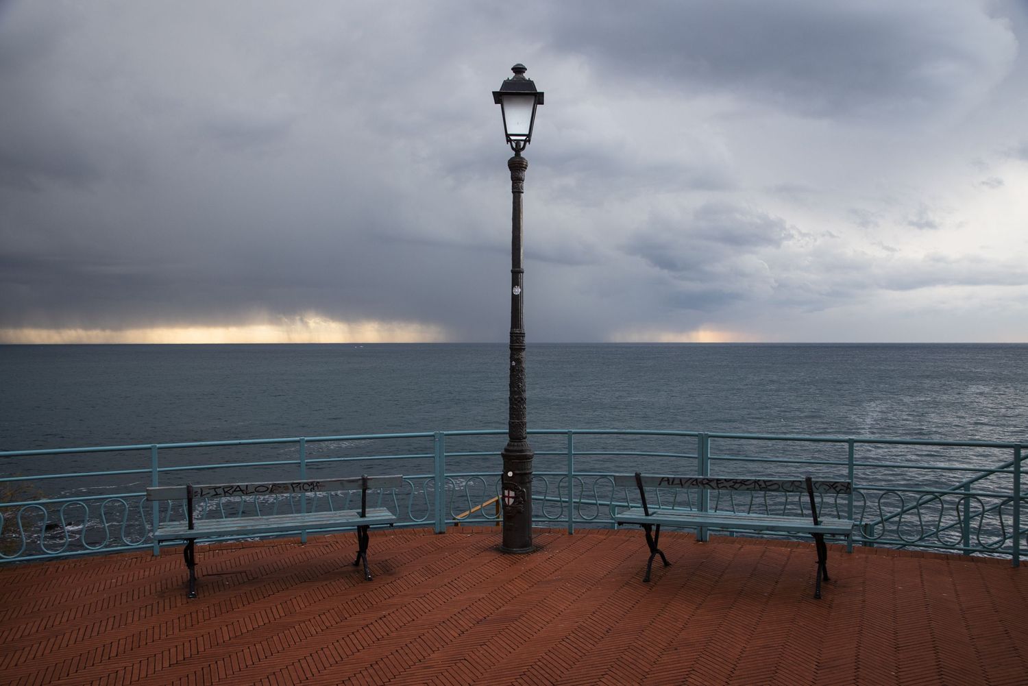 A stormy seaside promenade with two benches, a vintage lamp post, and dramatic cloudy skies over the ocean.
