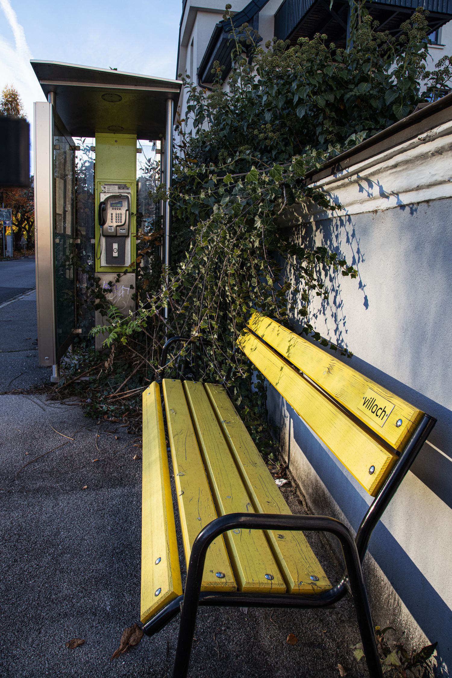 Yellow wooden bench beside a public telephone booth with ivy-covered wall in an urban street setting.