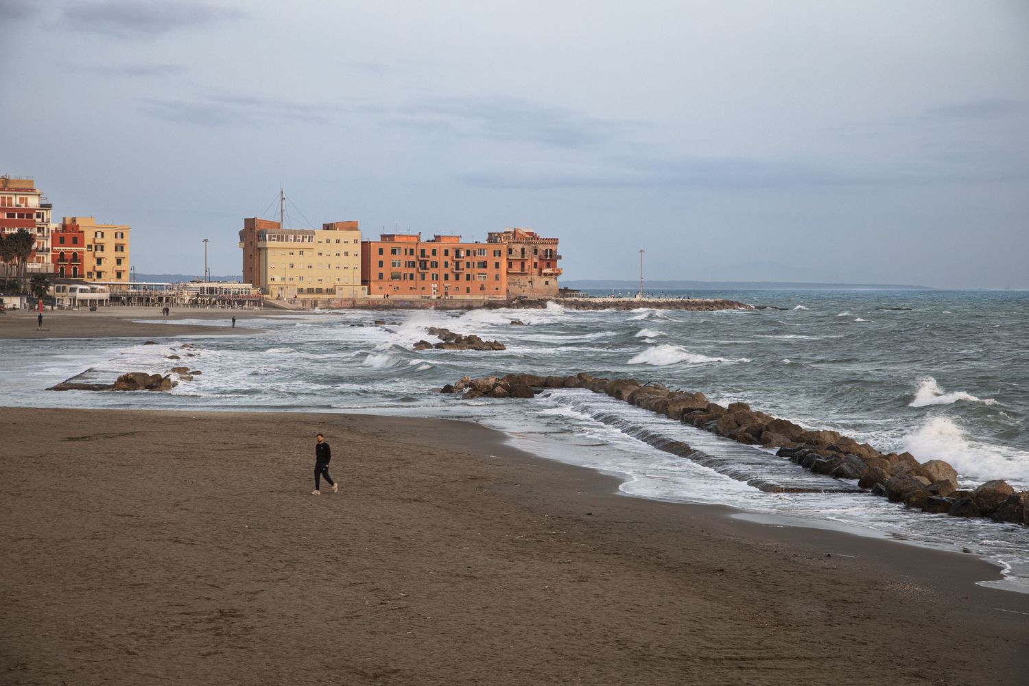 A lone person walks on a sandy beach with rough waves and orange buildings on a rocky coastline under cloudy skies.