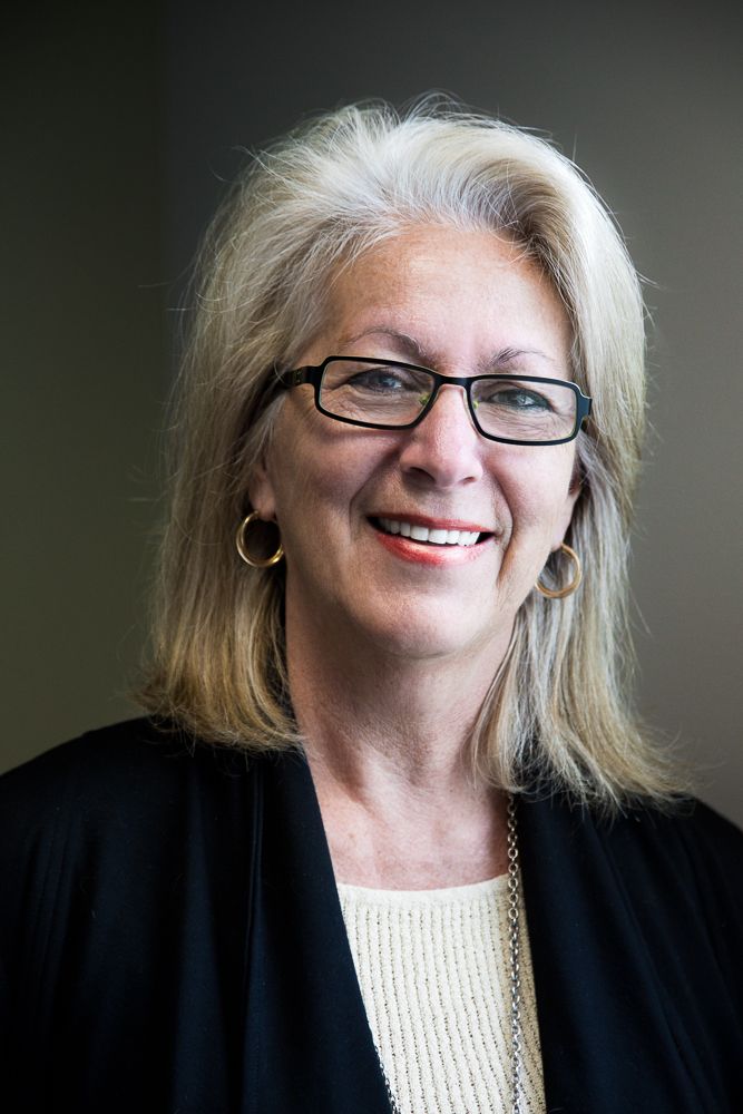 Professional portrait of someone wearing glasses and a black cardigan over a light sweater against a blurred background.