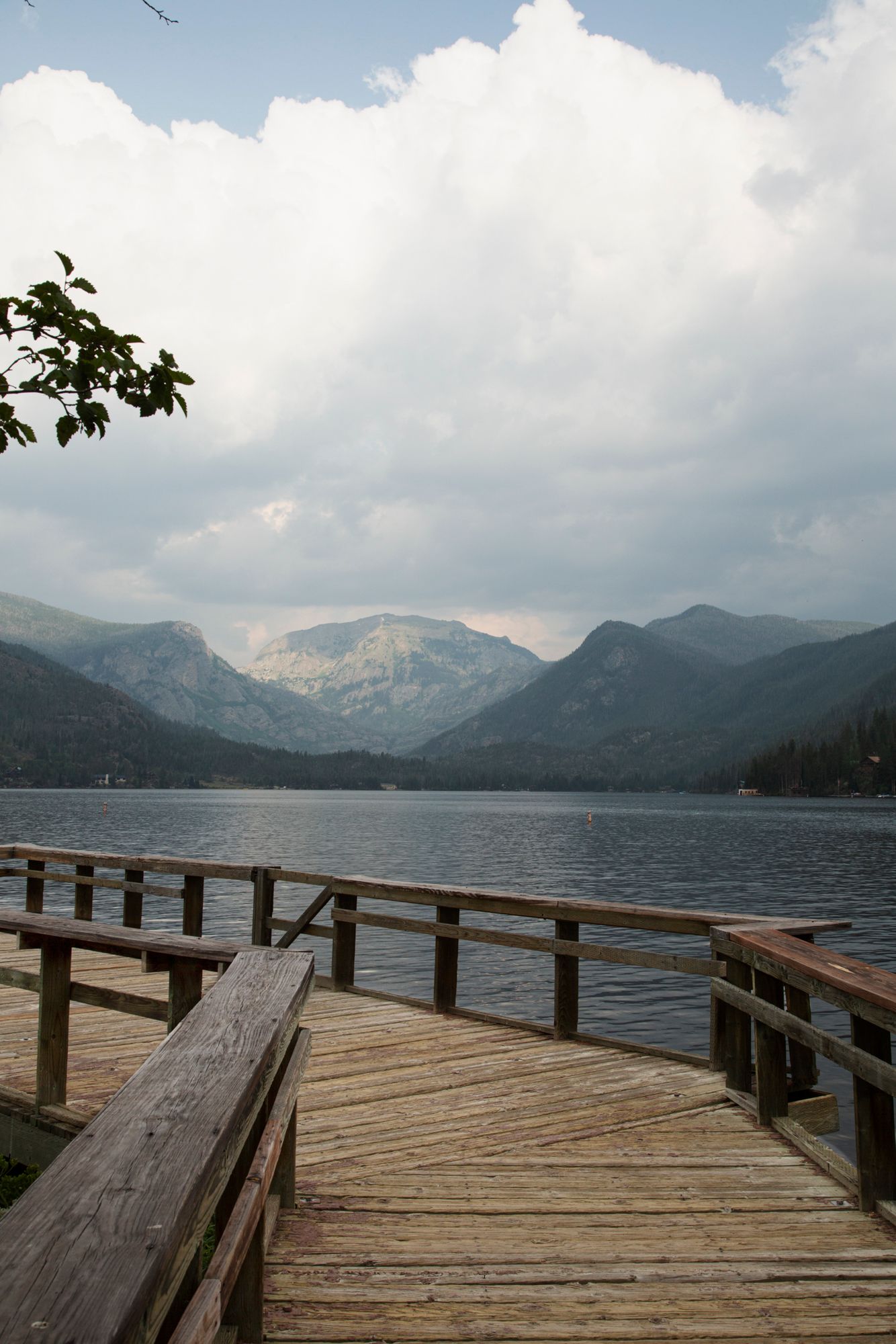 A panoramic view of a wooden dock extending into a mountain lake with cloudy skies and rugged peaks in the distance.