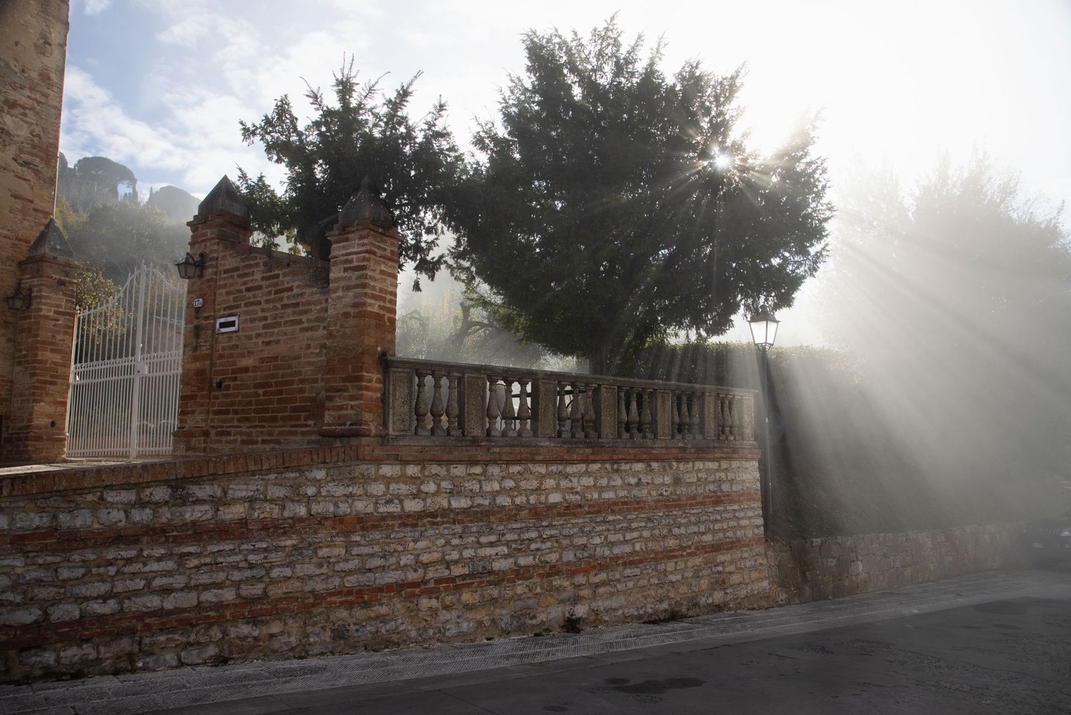Sunlight rays streaming through trees over an old brick wall and ornate gate in a historic outdoor setting.