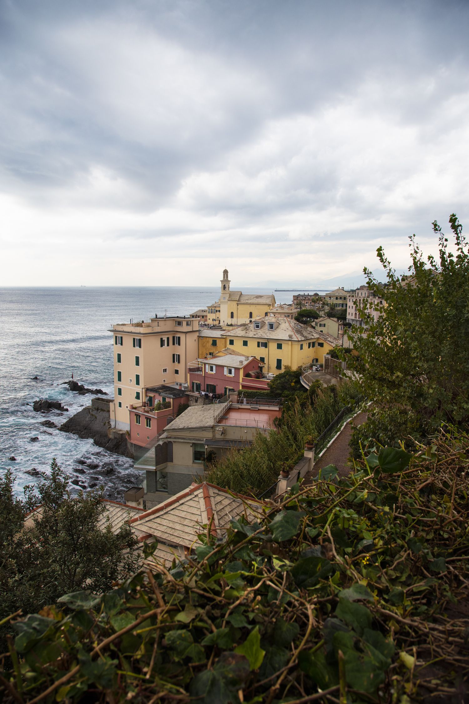 Colorful coastal village with historic buildings and a church tower overlooking the rocky Mediterranean sea under cloudy skies.
