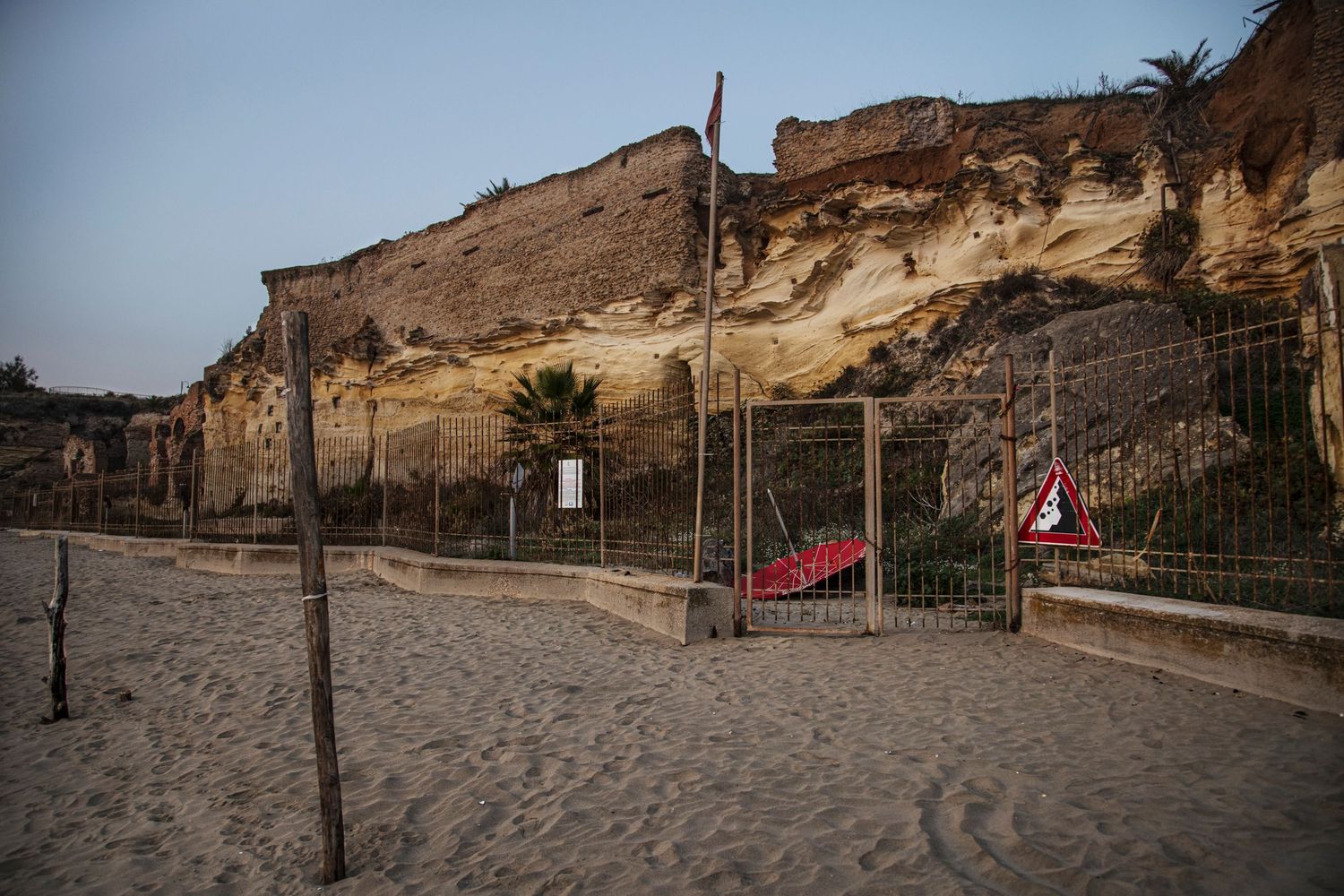 Sandy beach with fenced-off eroded cliff face and warning signs at dusk, showing coastal erosion hazard zone.