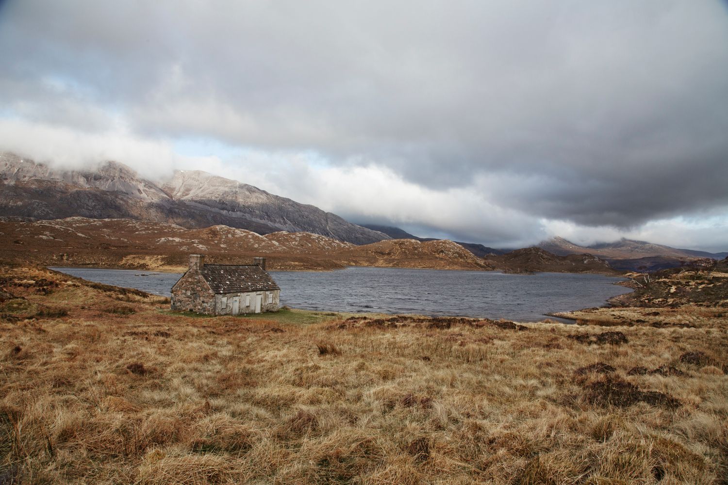 A rugged Scottish Highland landscape with a small loch and snow-dusted mountains under dramatic cloudy skies.