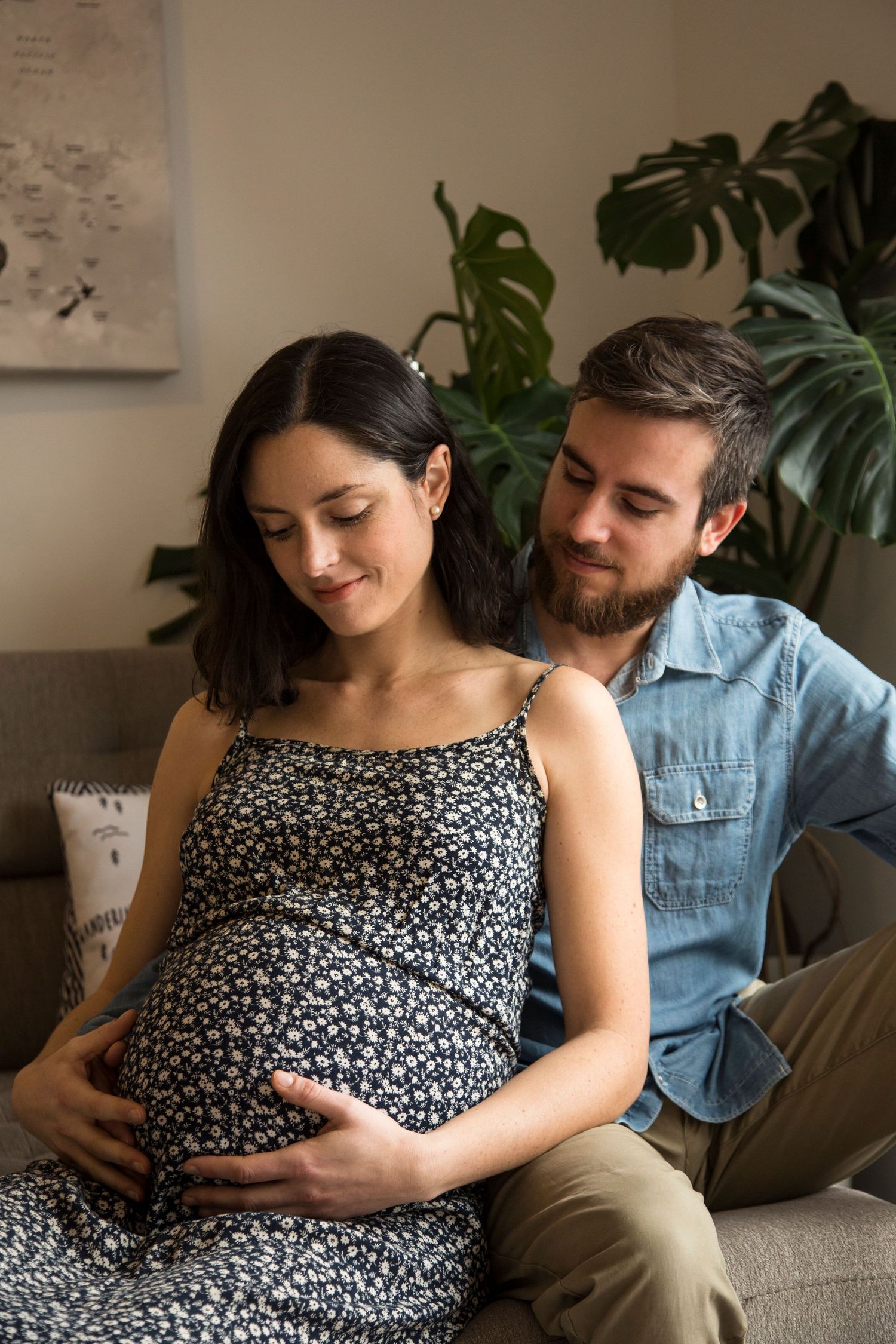 Indoor maternity photos with couple on couch surrounded by houseplants.