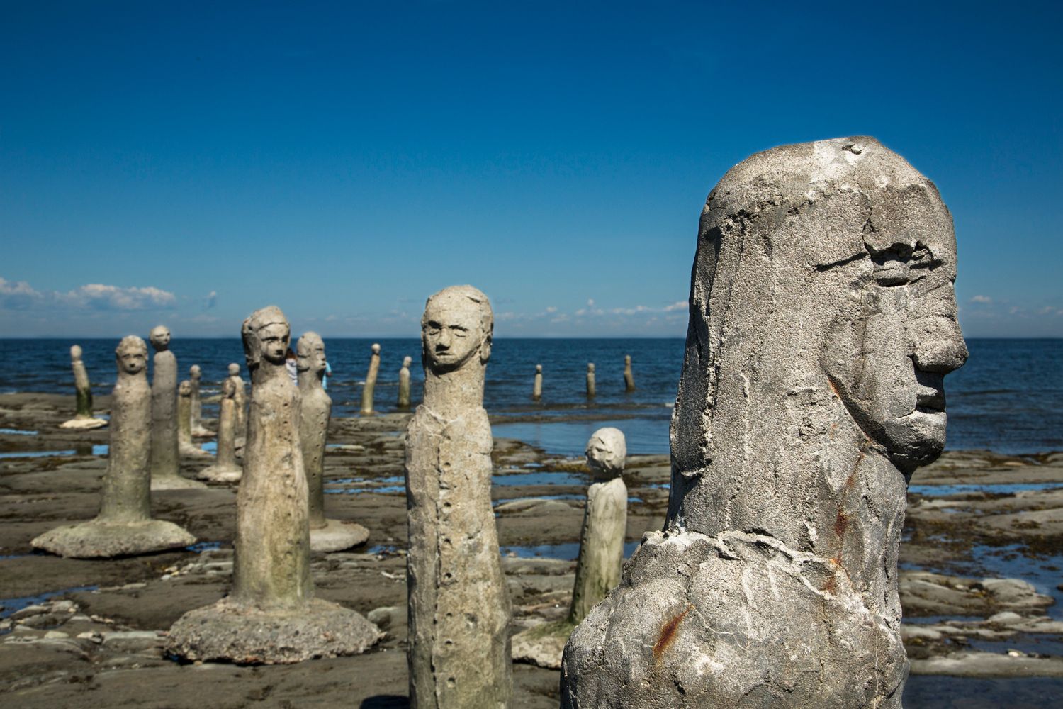 Ancient stone sculptures line the rocky shore against a bright blue ocean backdrop.