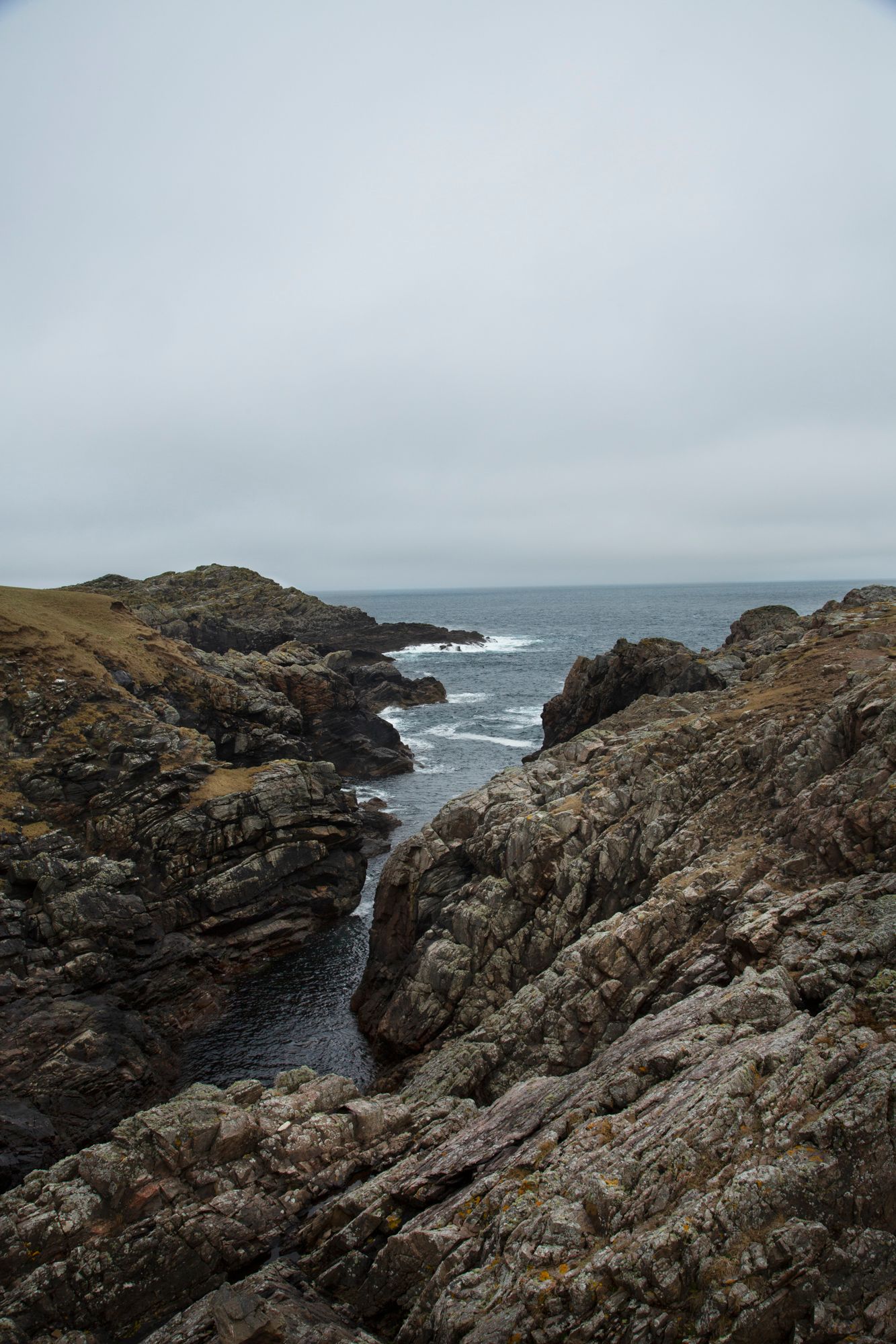 Rocky coastal landscape with rugged cliffs and crashing waves against the shoreline under an overcast sky.