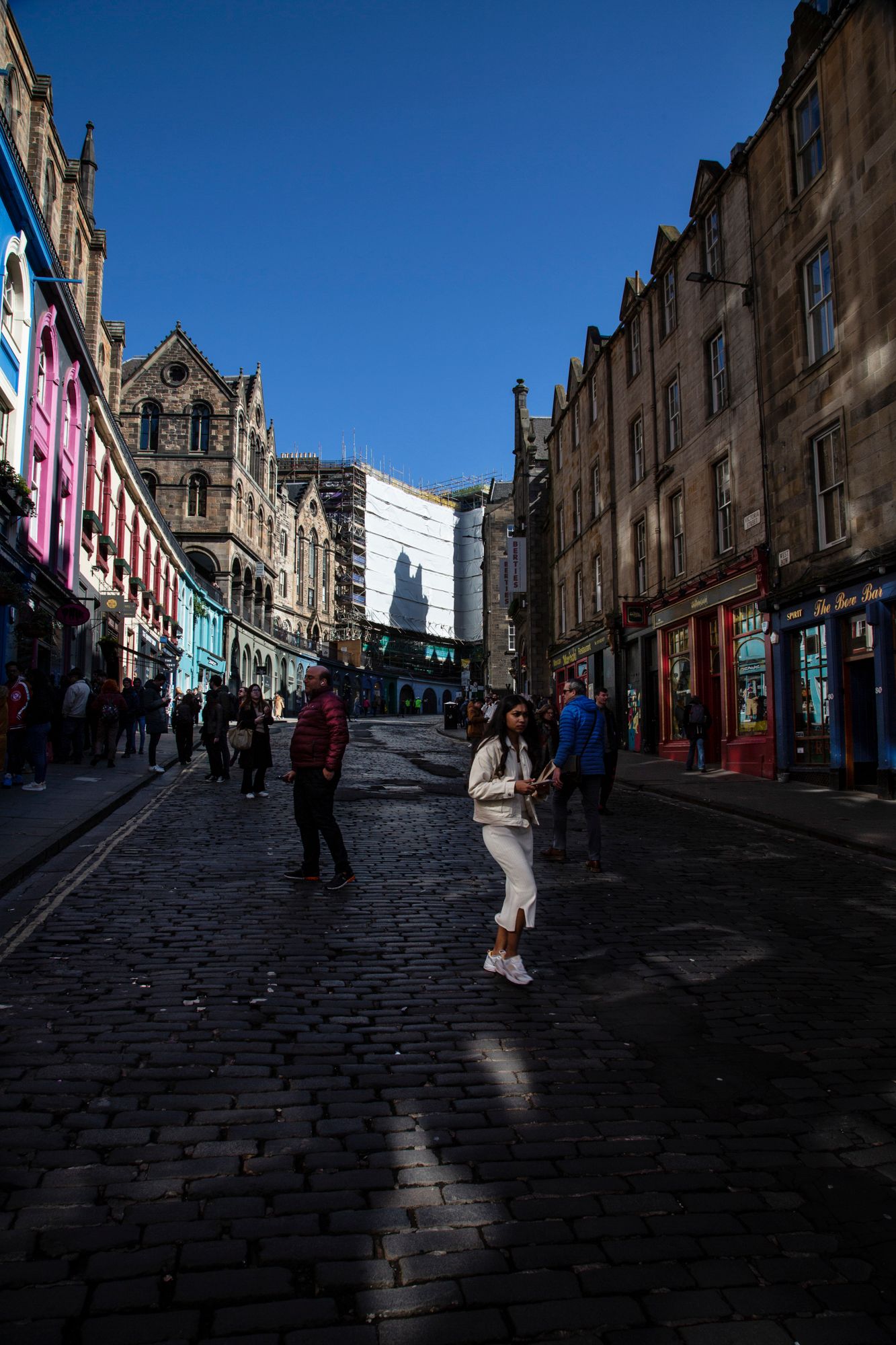 Cobblestone street in Edinburgh's historic Royal Mile with pedestrians walking between old stone buildings in shadow.