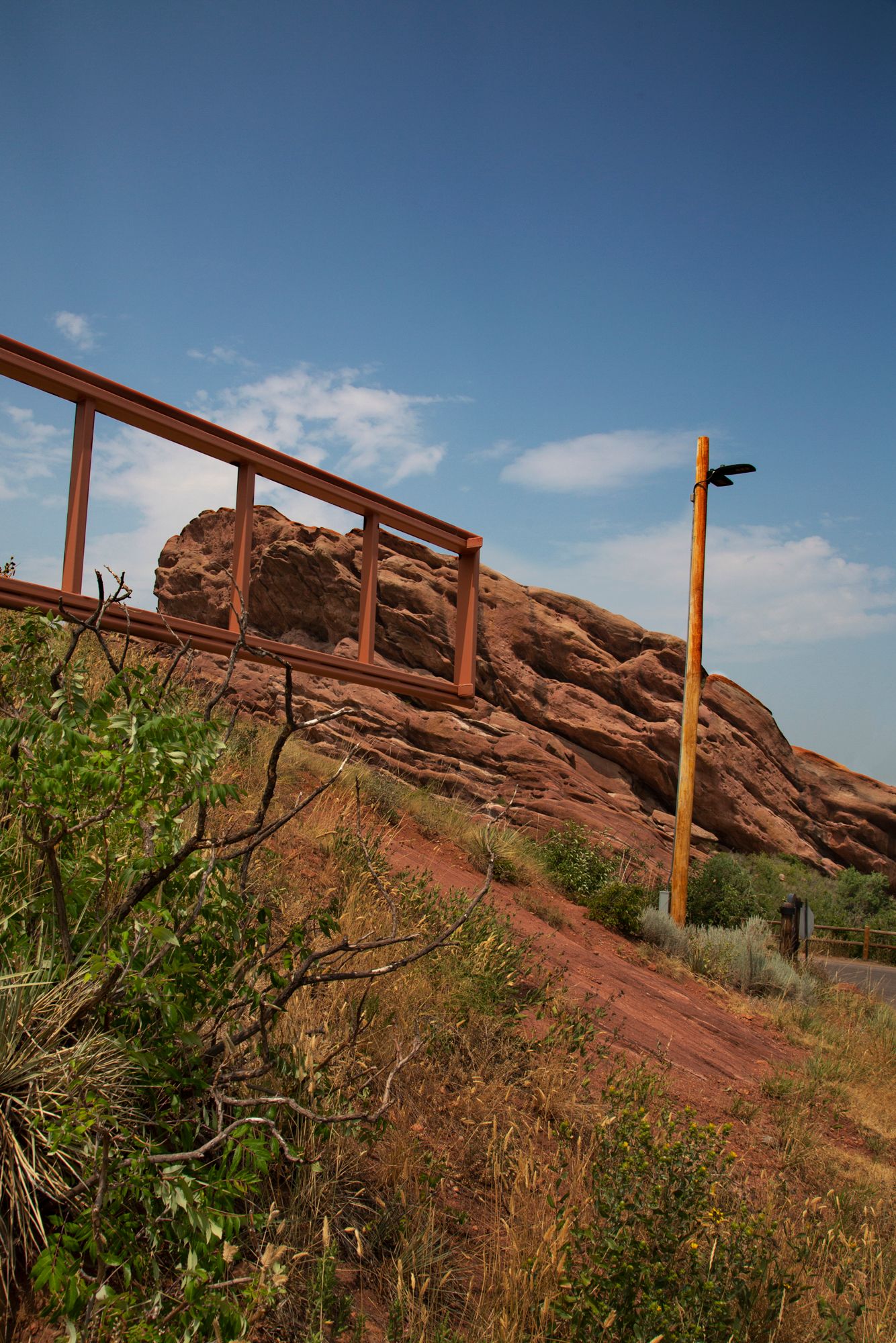A rustic railing and lamp post stand against red rock formations at Red Rocks Park in Colorado under blue skies.