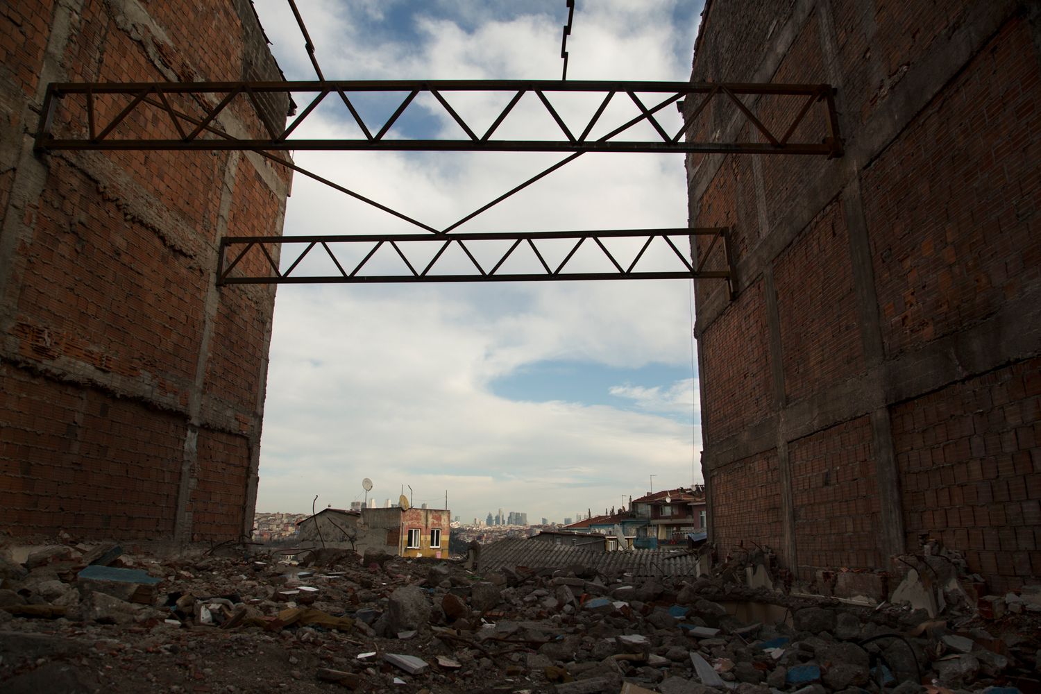 Industrial metal truss structure spans between brick walls against cloudy sky.