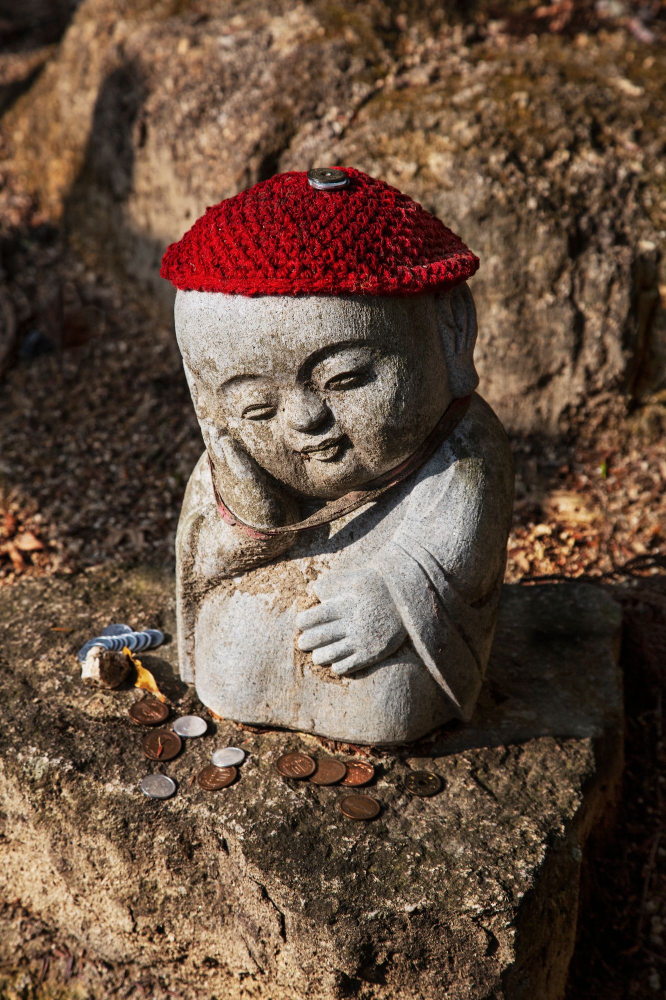Small stone statue wearing a red knit cap sits on rocky surface in traditional Japanese style.