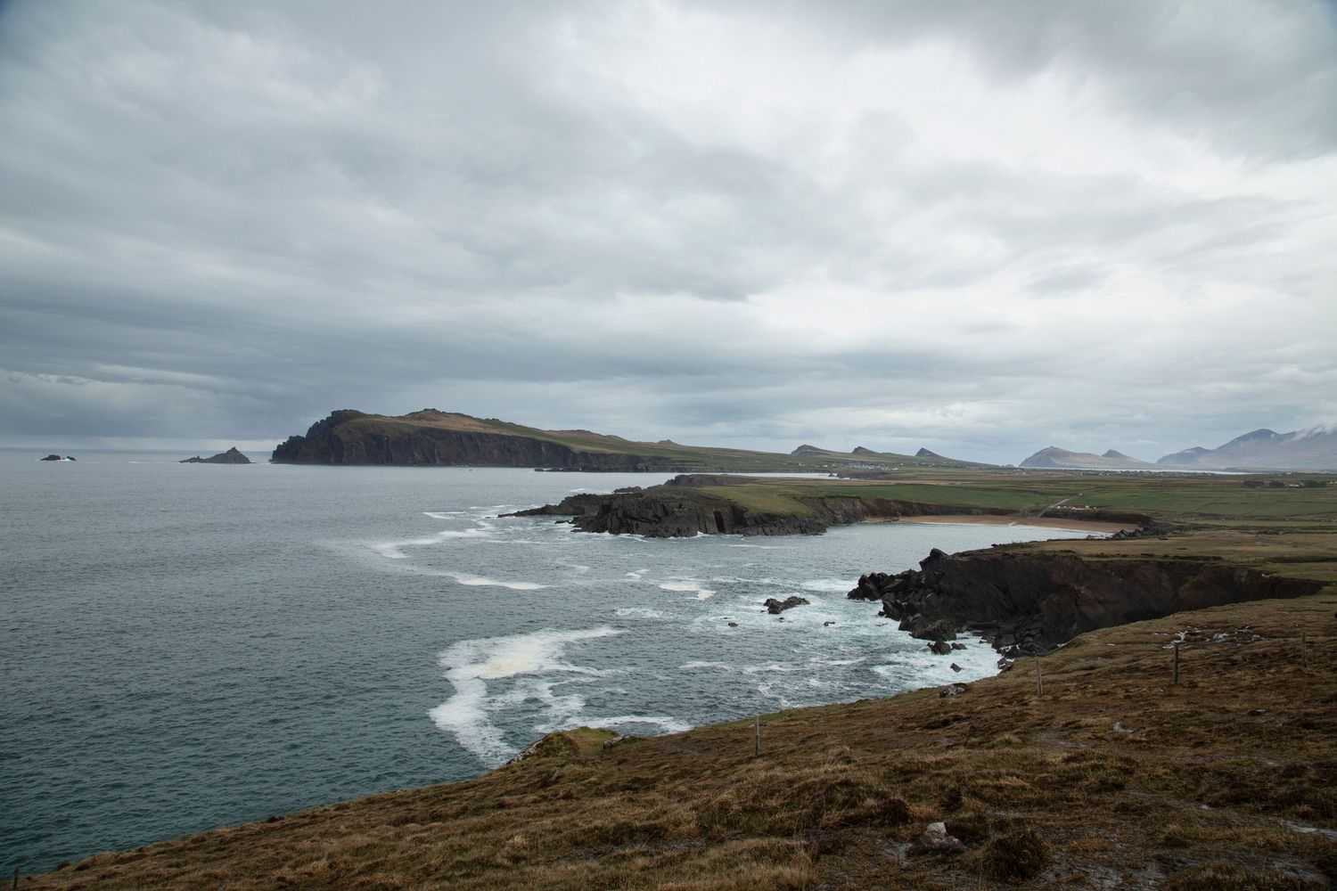 Dramatic coastal landscape with rocky cliffs and crashing waves under moody skies along a rugged shoreline.