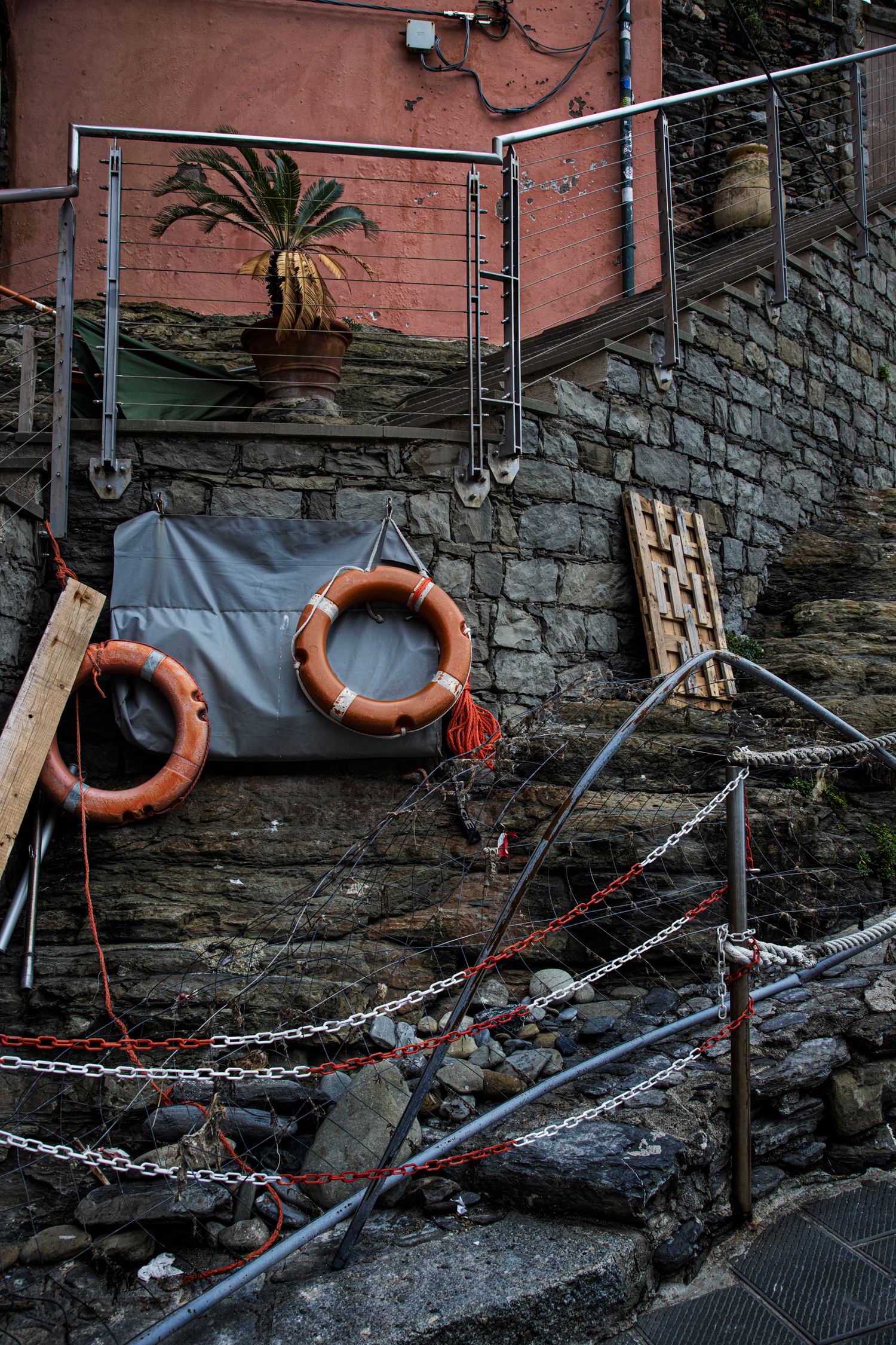 Orange life rings hang on a stone wall near metal railings and chains at a coastal dock in Cinque Terre, Italy.