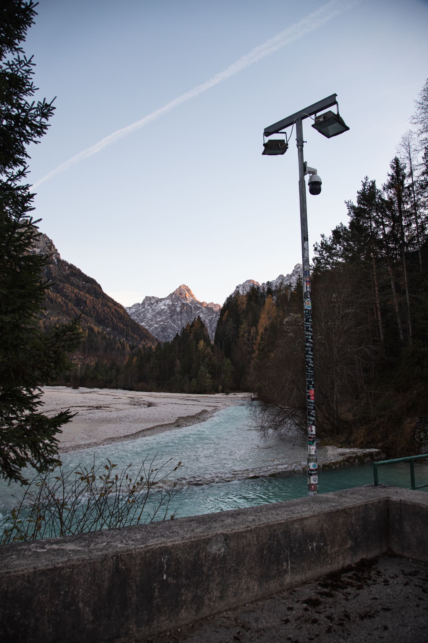 A street lamp stands beside a turquoise alpine river with snow-capped mountains and dense forests in the background at dusk.