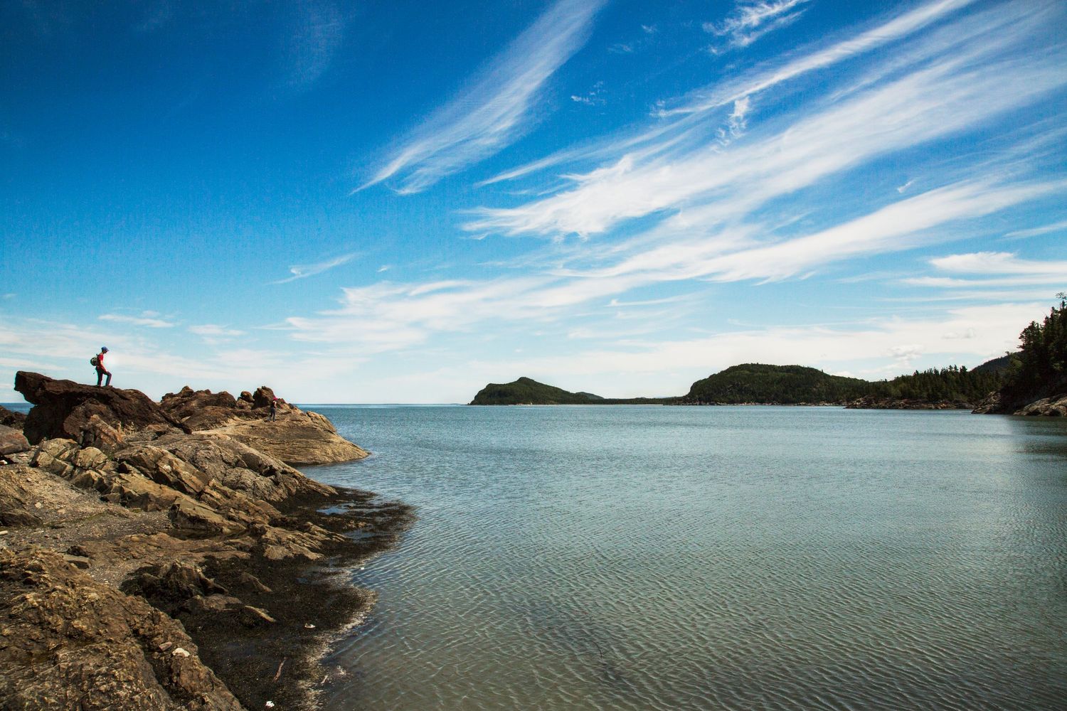 A silhouetted figure stands on rocky coastal cliffs overlooking a tranquil bay with wispy clouds in a bright blue sky.