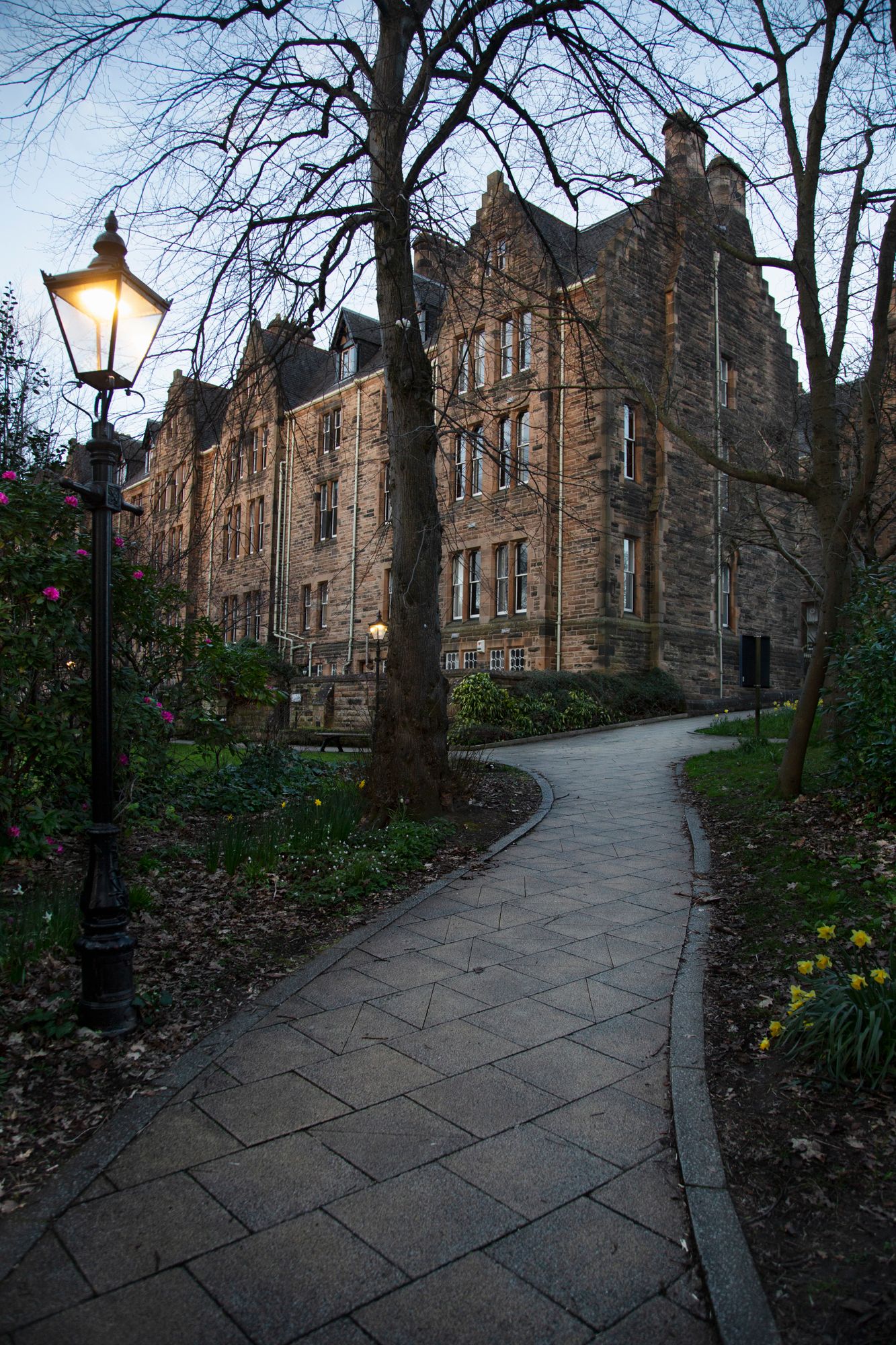A curved stone path leads through historic Edinburgh buildings at dusk with a glowing lamppost illuminating the scene.