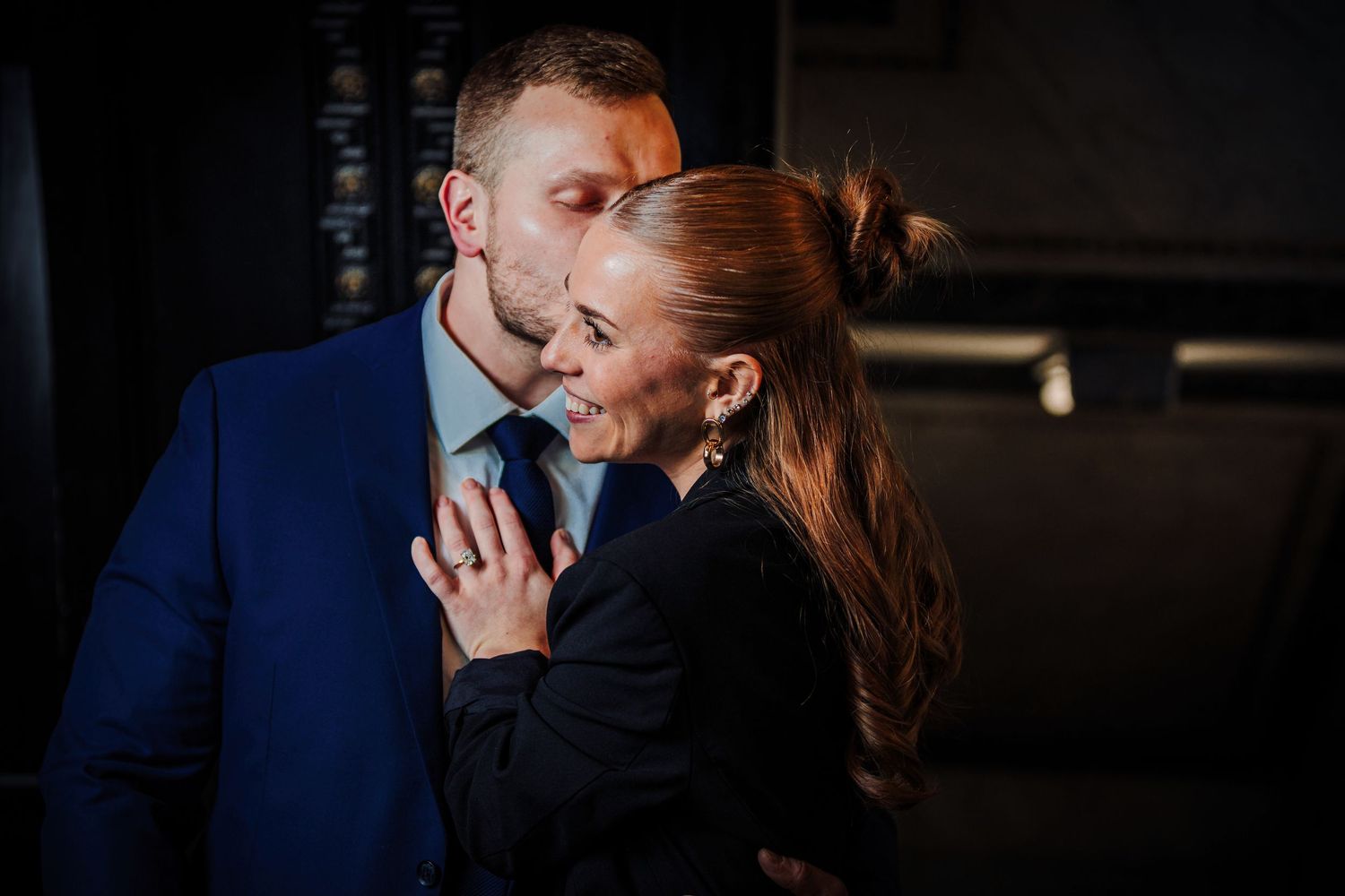 A romantic couple embraces in formal attire, with the woman in black and the man in a navy blue suit.