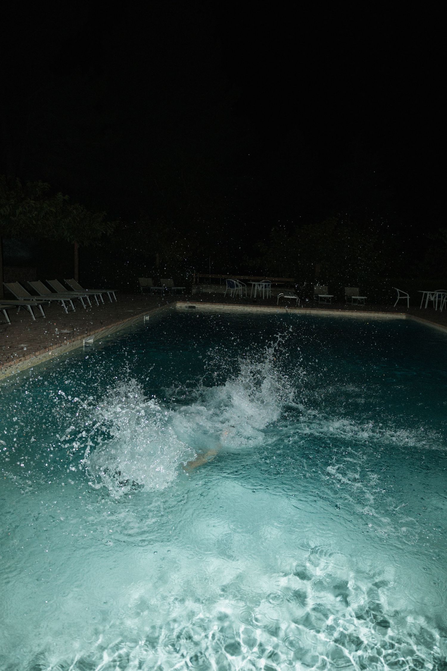 A dramatic splash erupts in an illuminated swimming pool at night with lounge chairs visible around the edge.
