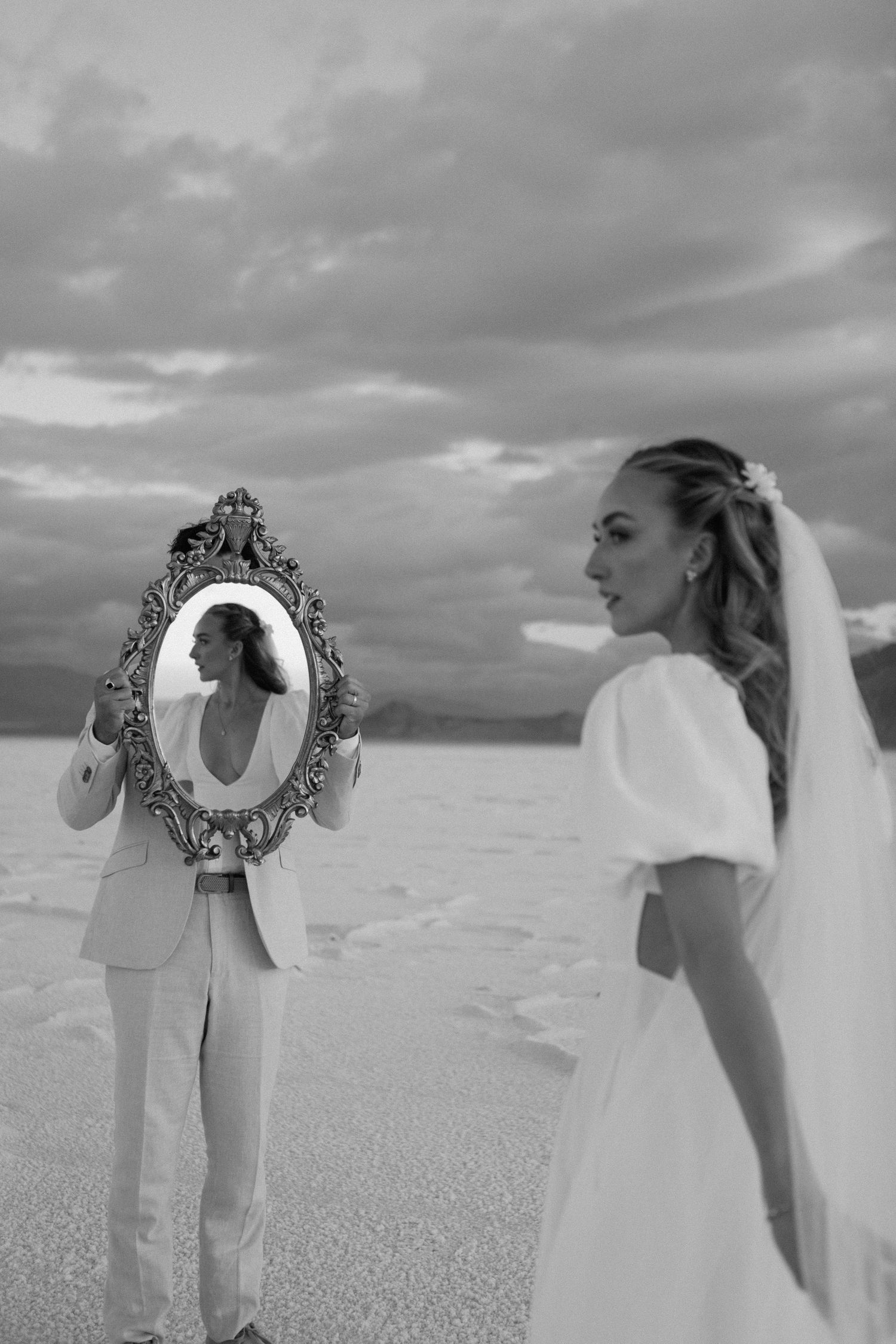 Two figures on salt flats, one in a wedding dress and veil while another holds an ornate mirror in dramatic black and white.