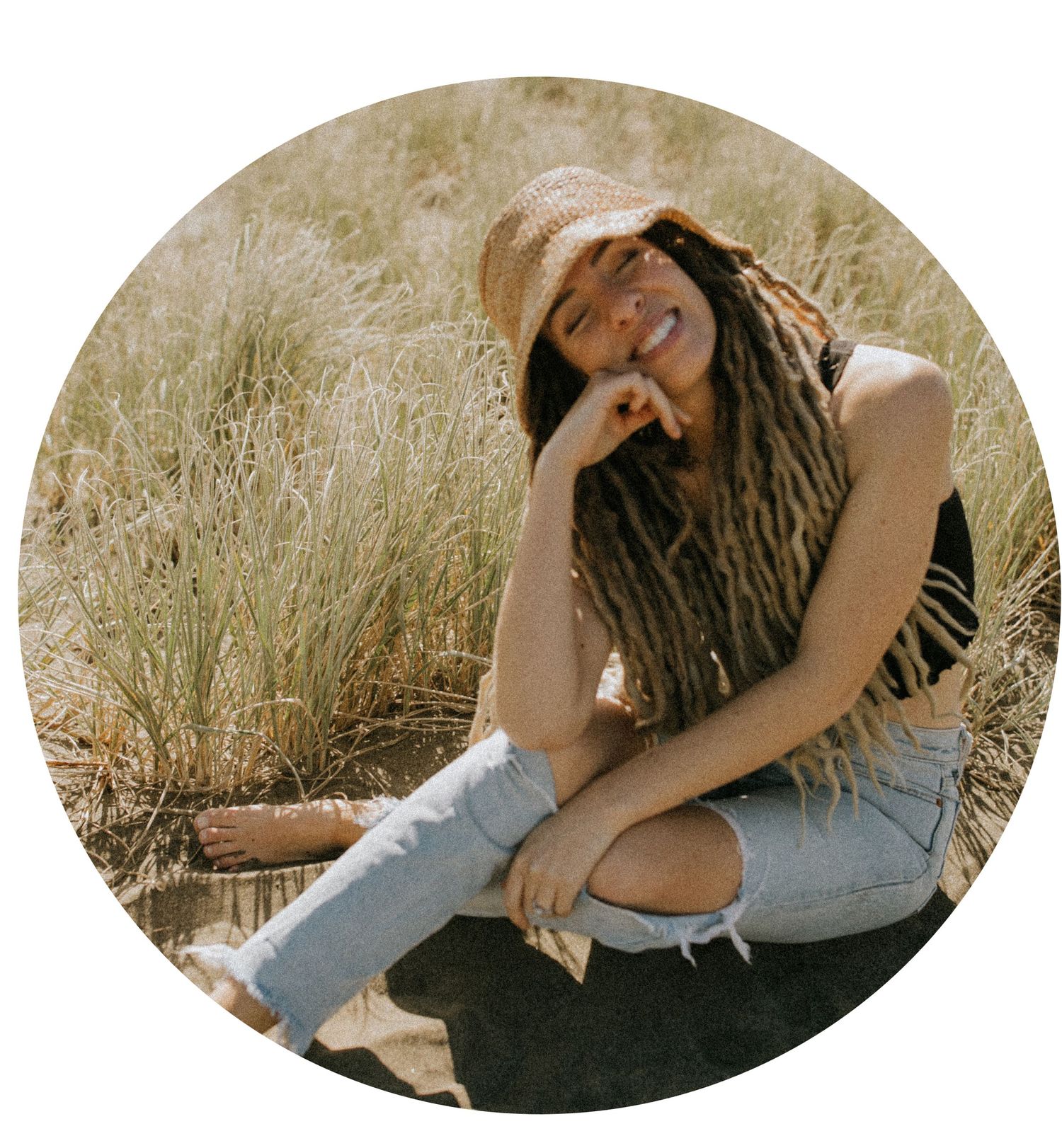 A person sitting in a sunlit field wearing distressed denim jeans and dreadlocks, laughing candidly.