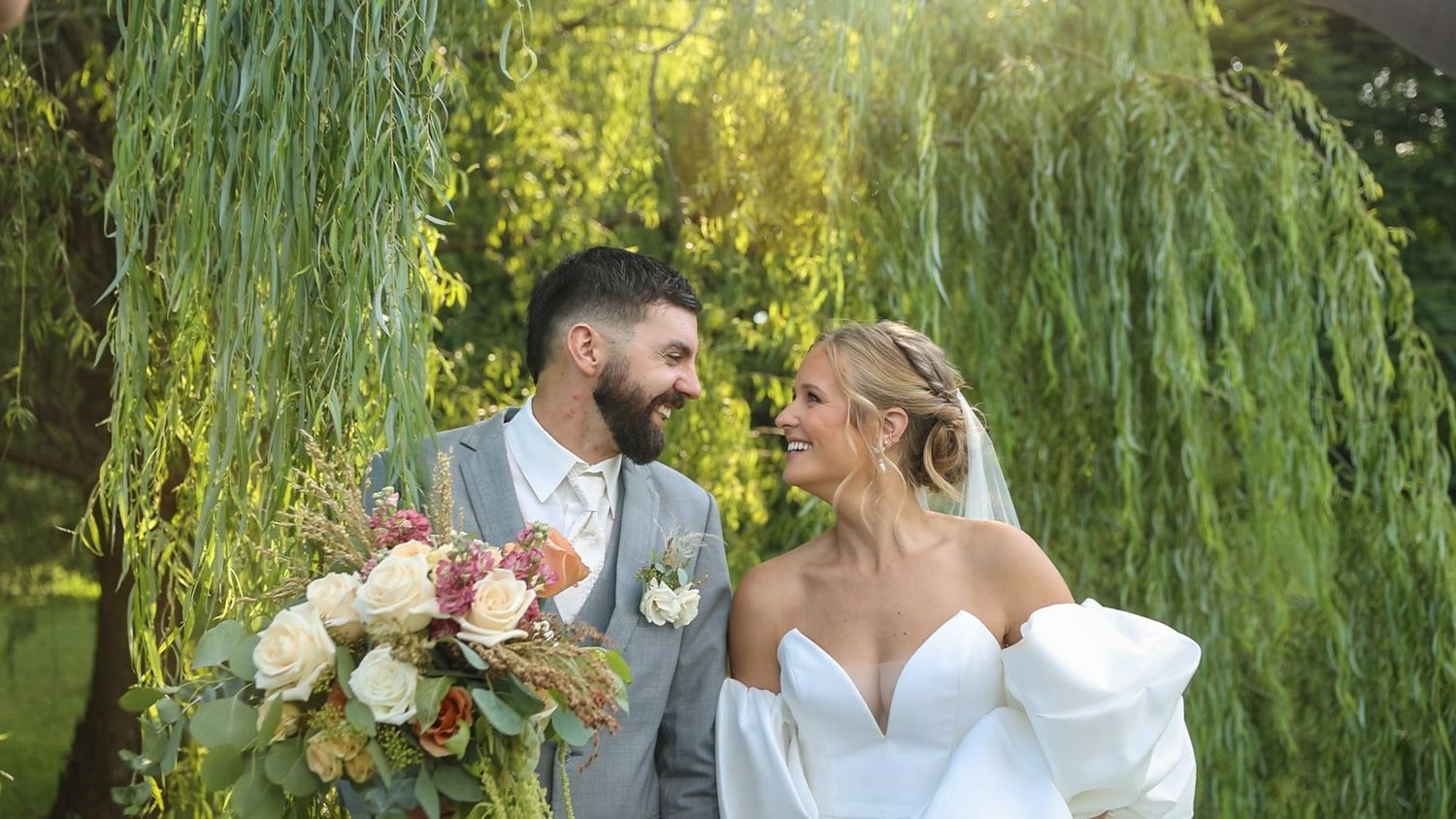 Elegant wedding couple stands together under weeping willow tree with delicate pink and white floral bouquet.