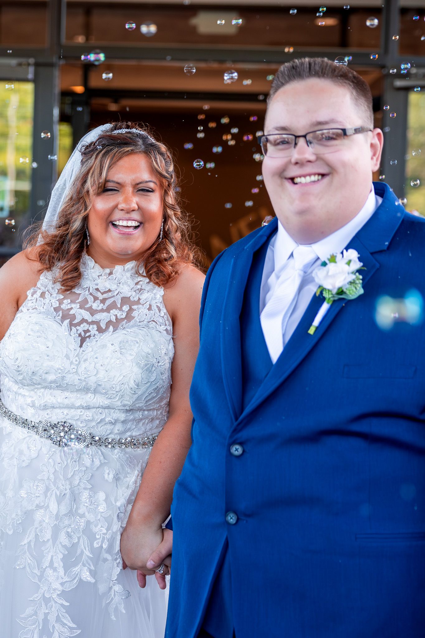 A happy couple on their wedding day posing together, the bride in lace and the groom in a blue suit with bubbles floating around.