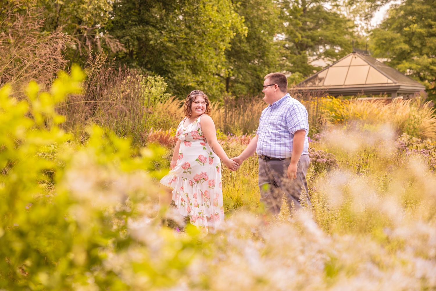 A couple walks hand in hand through a sunlit meadow with tall grass and wildflowers during golden hour.
