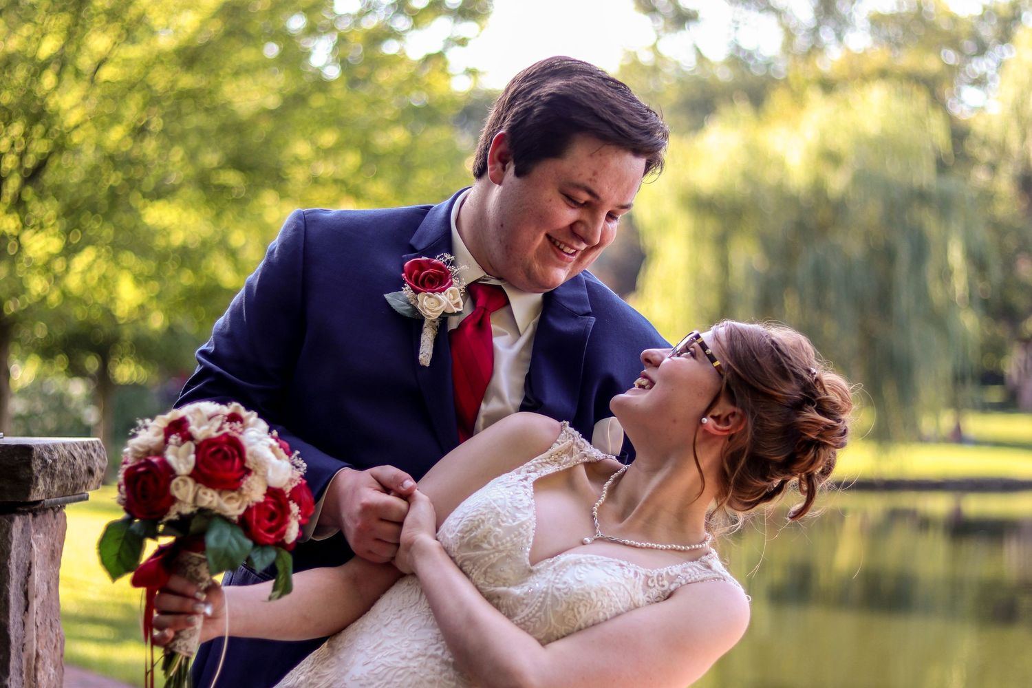A joyful wedding couple shares a candid moment outdoors near a pond with a beautiful red and white bouquet.