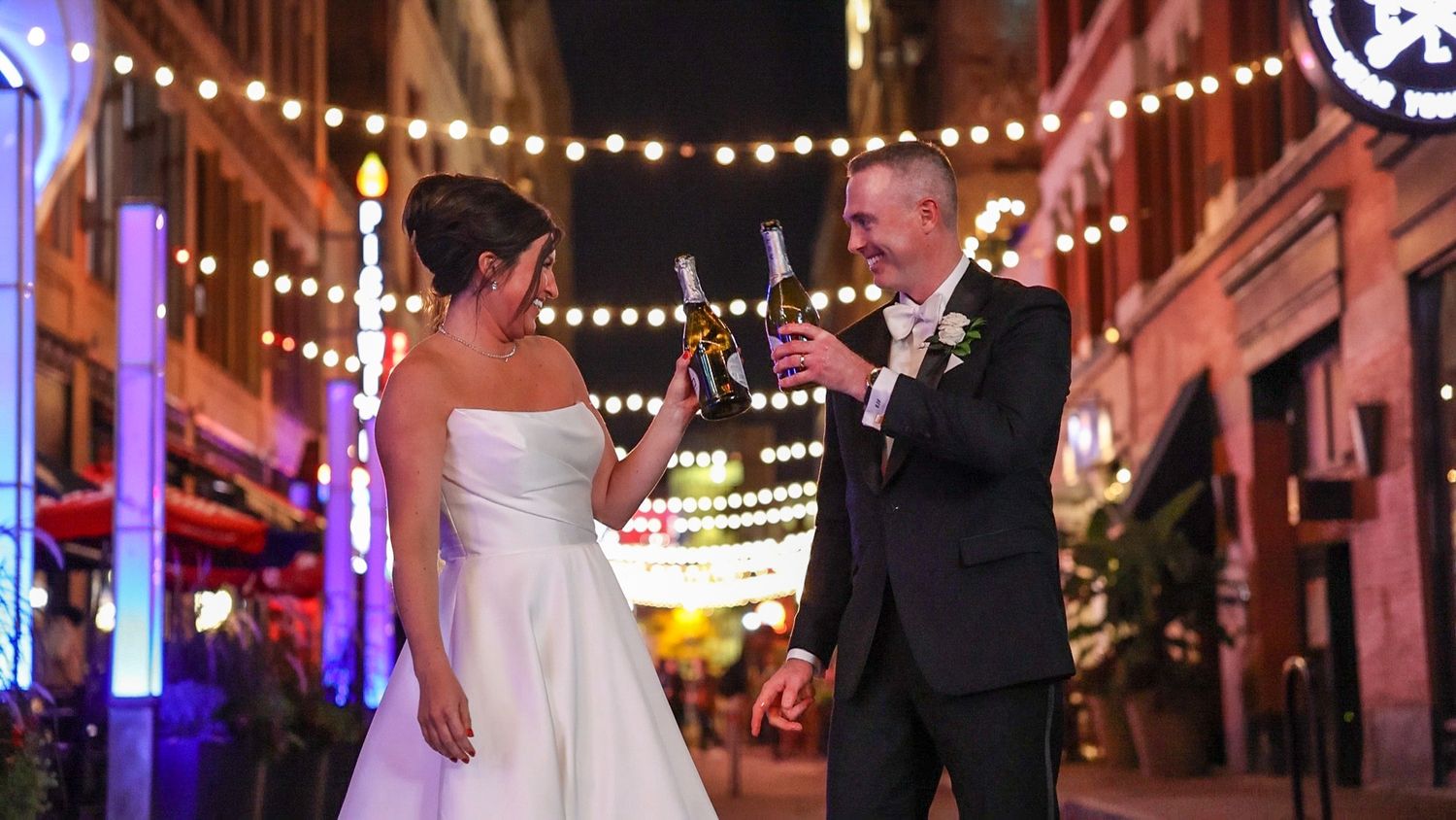 A couple stands under glowing string lights on a festive urban street at nighttime during a celebration.