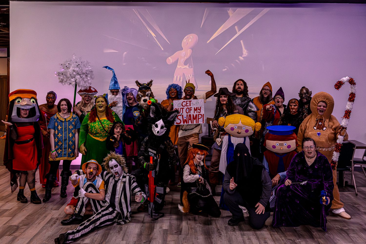 Large group in Halloween costumes posing together against purple wall with decorations.