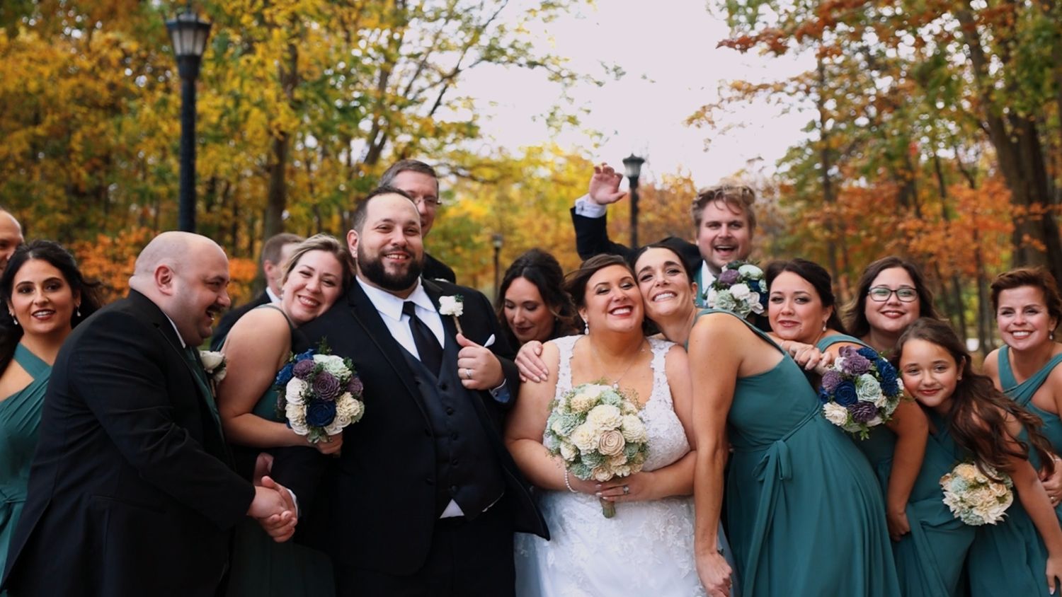 Wedding party in formal attire laughs together during autumn photoshoot with colorful fall foliage in background.