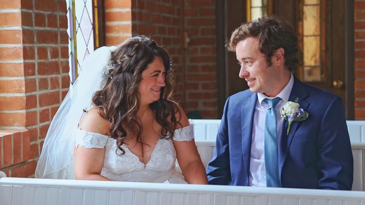 Bride in white off-shoulder dress and groom in navy suit share a joyful moment together on their wedding day outside church.