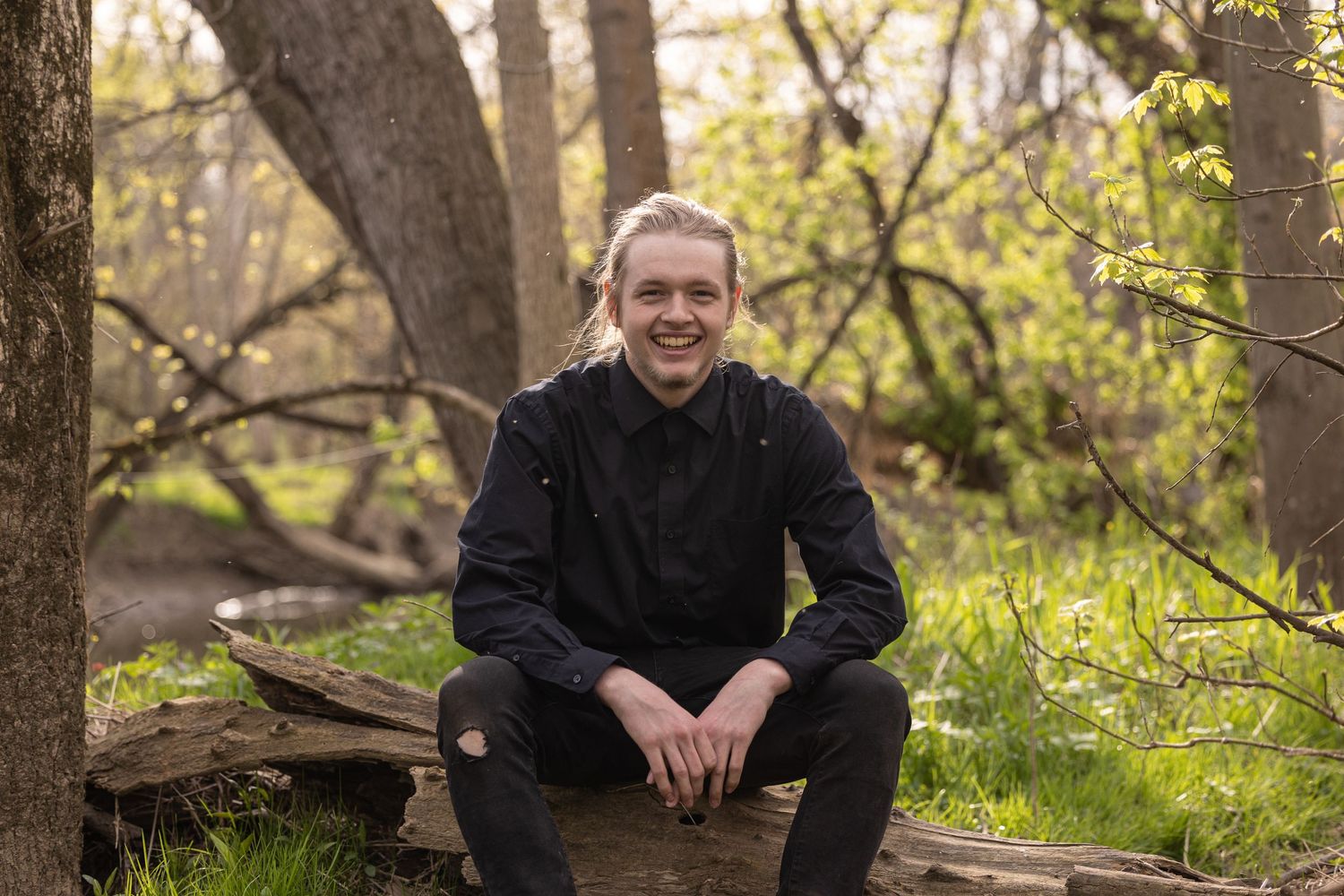Portrait of a person in a black sweater sitting on a wooden log in a sunlit forest setting.