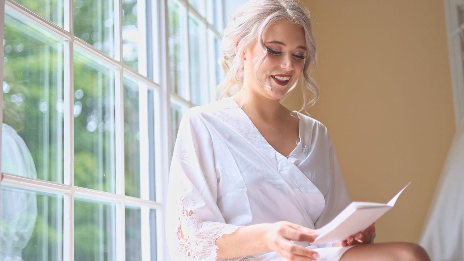 Someone in a white robe reads by sunlit windows while smiling warmly on a bright morning.