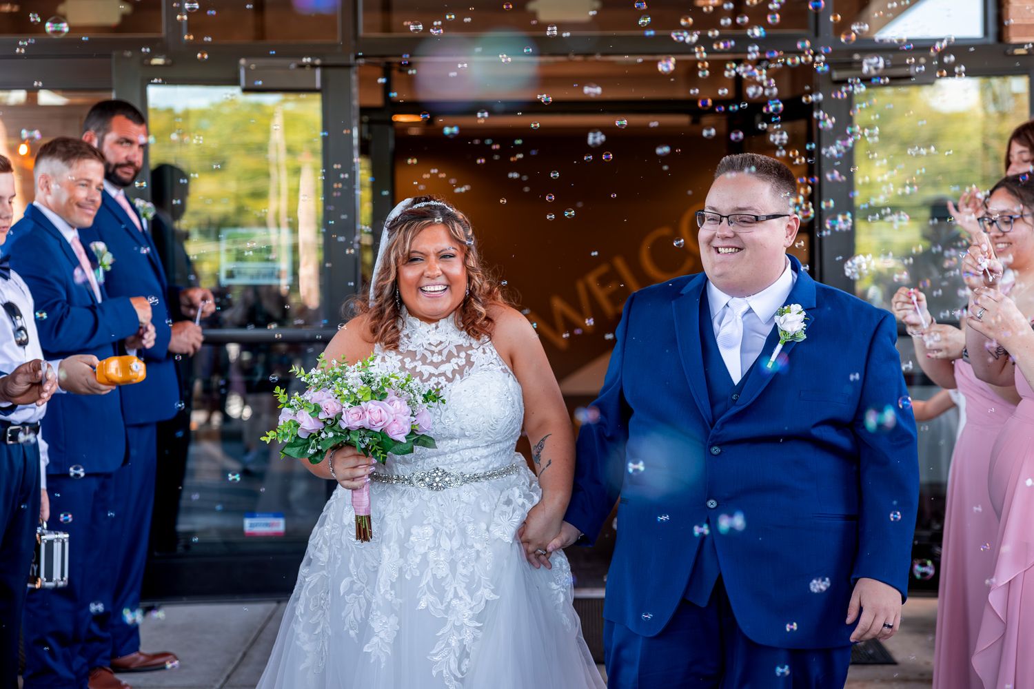 A wedding couple exits while surrounded by bubbles on their special day wearing white and navy blue attire.