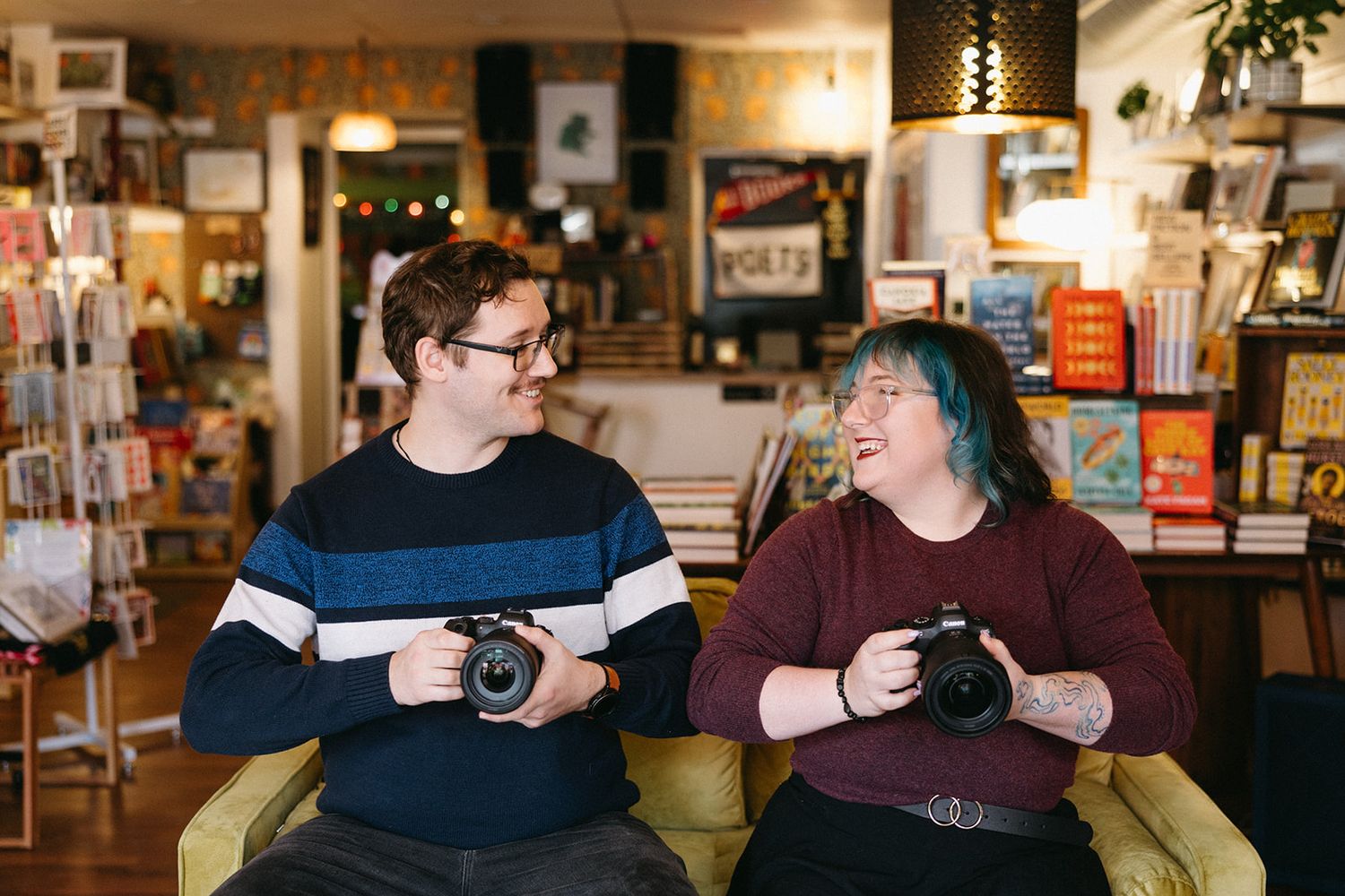 Two photographers sit together holding cameras in a cozy bookstore setting.