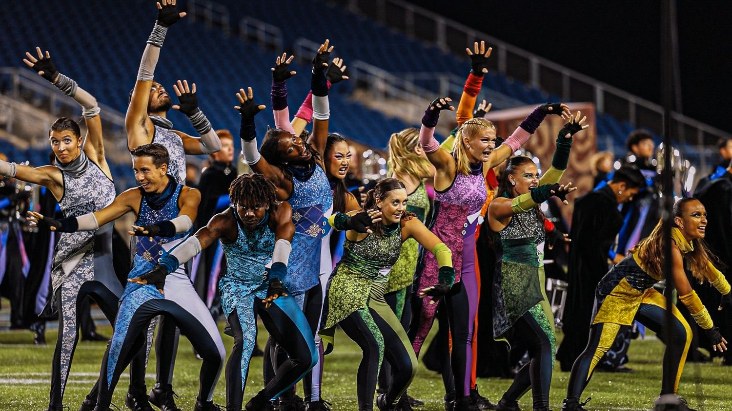 Group of performers in colorful costumes performing synchronized dance moves in stadium.