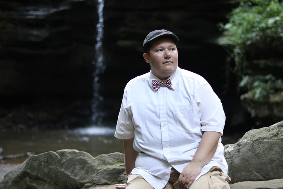 Someone wearing a white button-down shirt, bow tie and cap sits on rocks near a waterfall in nature.
