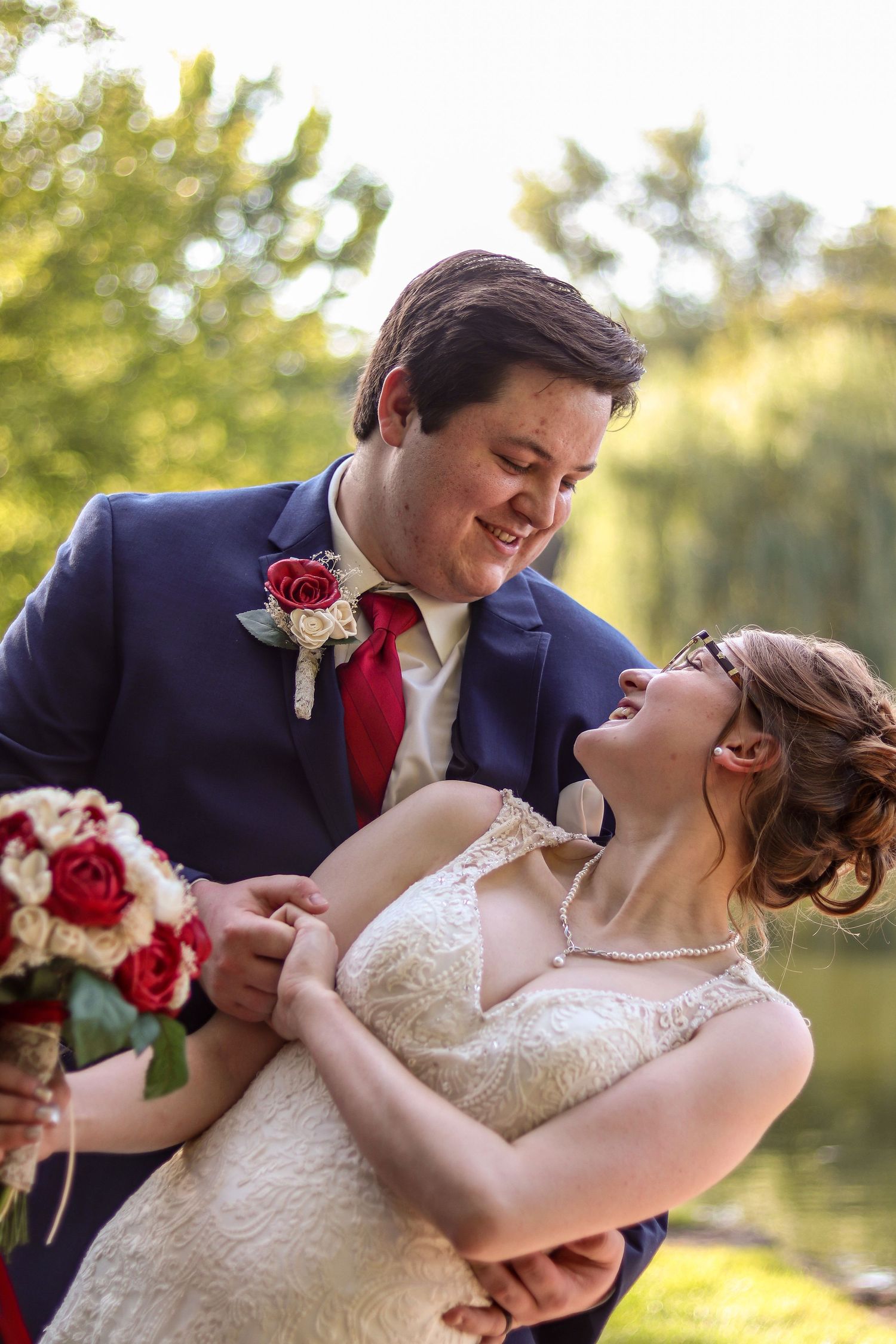 A romantic wedding photo outdoors with a groom in navy suit and red tie dipping bride in lace dress with red bouquet.