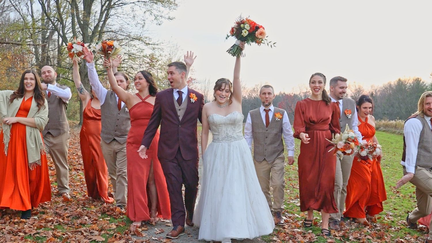 Wedding party in bright orange and white attire celebrates joyfully outdoors on an autumn day with raised bouquets.
