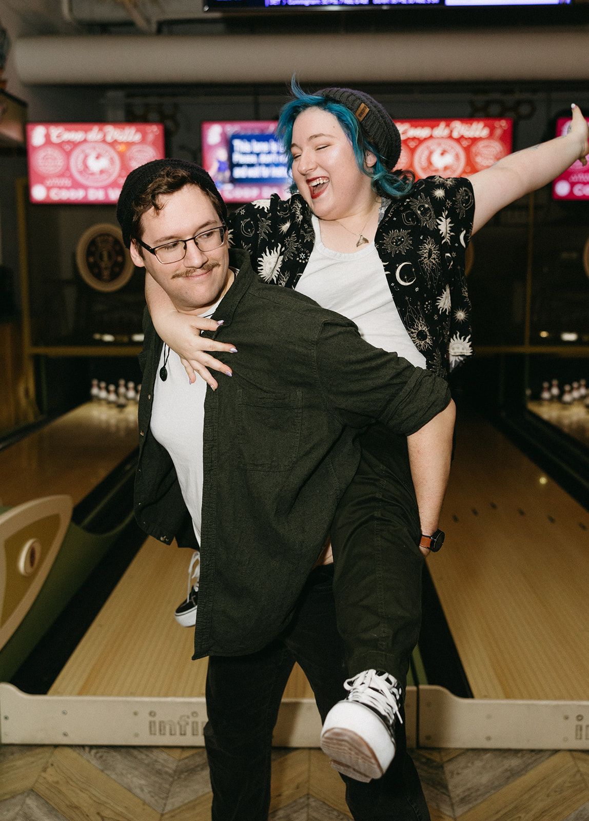 Two people share a playful moment at a bowling alley, one person carrying the other in a dramatic pose on the lanes.