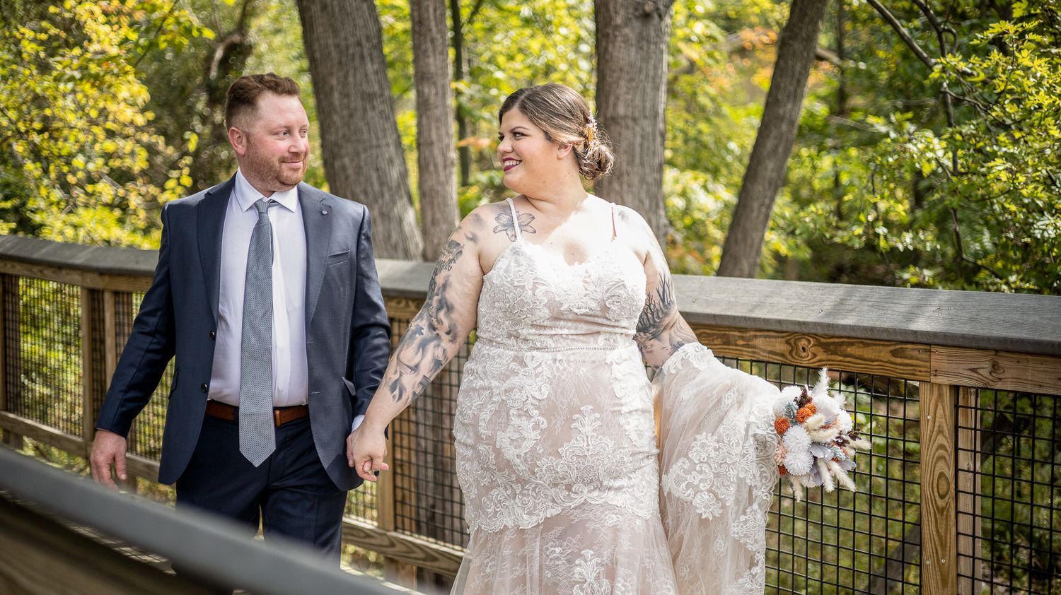 A couple in wedding attire walking together on a wooden bridge surrounded by autumn foliage.