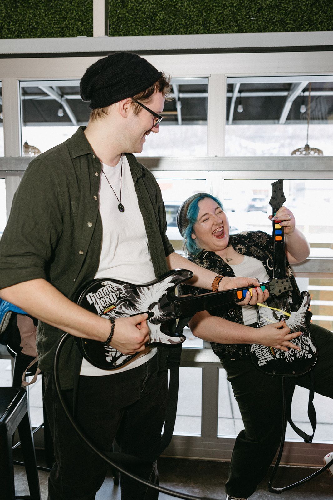 Two musicians play video game controllers as instruments against a white window backdrop during an indoor performance.