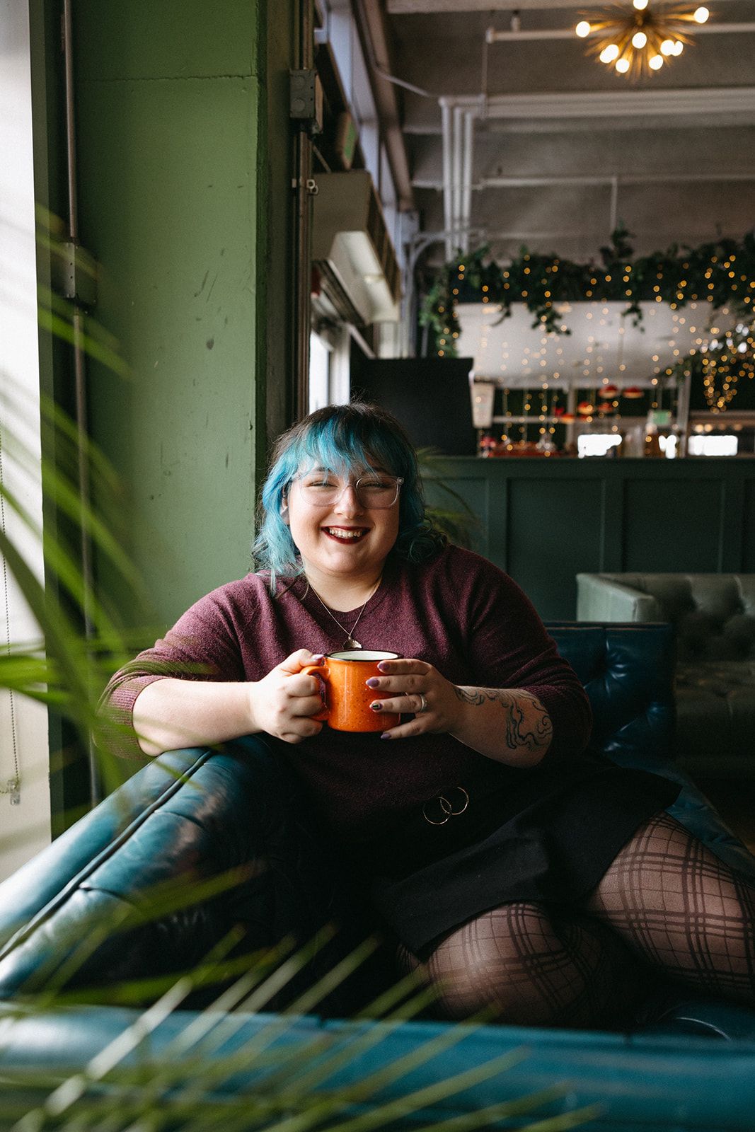 Person with blue hair sits comfortably at restaurant booth holding orange coffee mug near sunny window with plants.