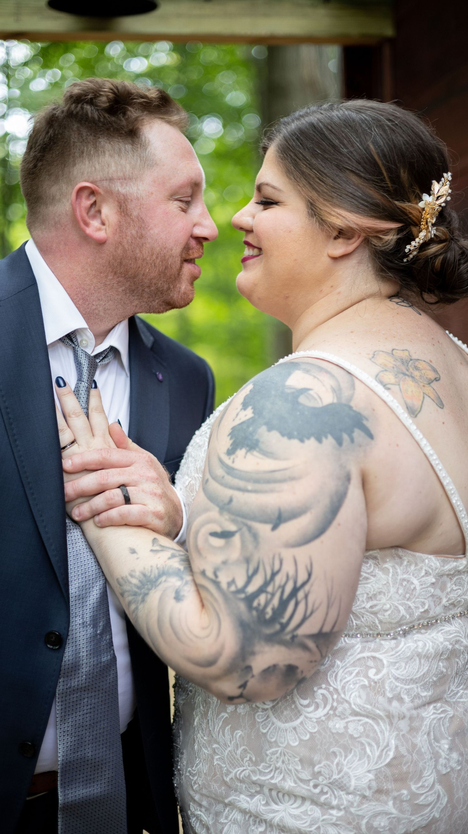 A happy couple share an intimate moment on their wedding day, the bride wearing a lace gown and displaying ornate arm tattoos.