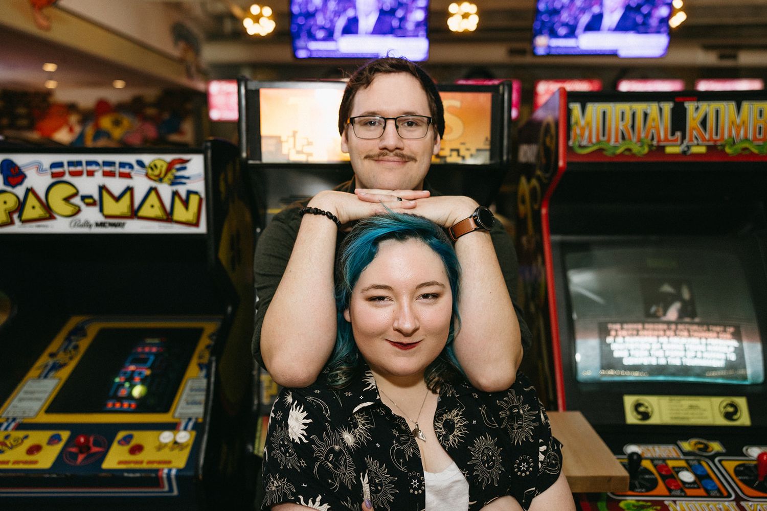 A couple poses playfully at a retro arcade with classic games like Pac-Man visible in the background.