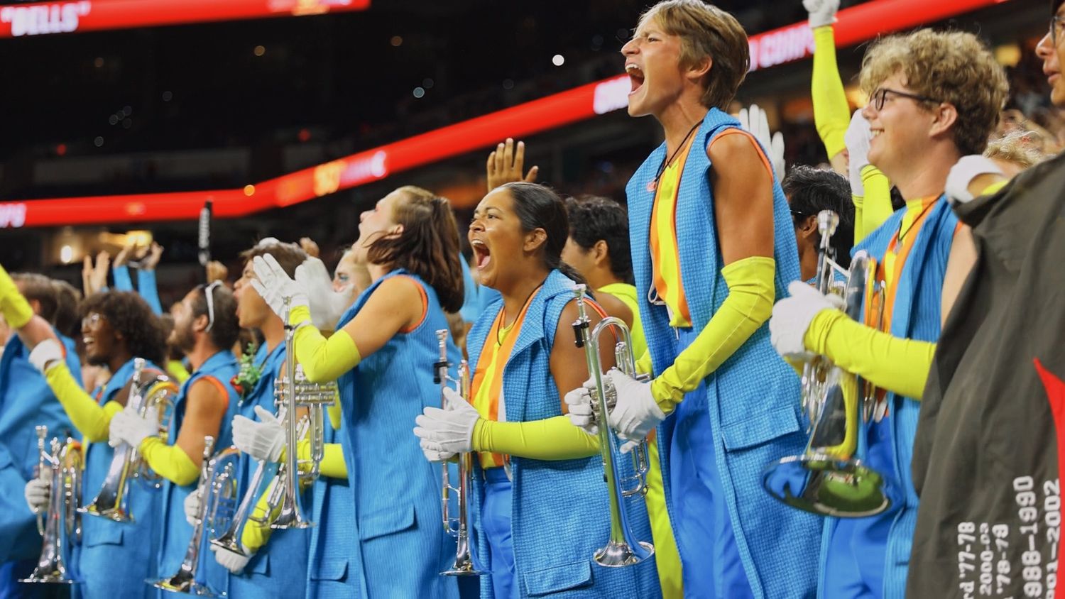 Basketball fans in bright blue and yellow uniforms cheer enthusiastically from the sidelines during a game.
