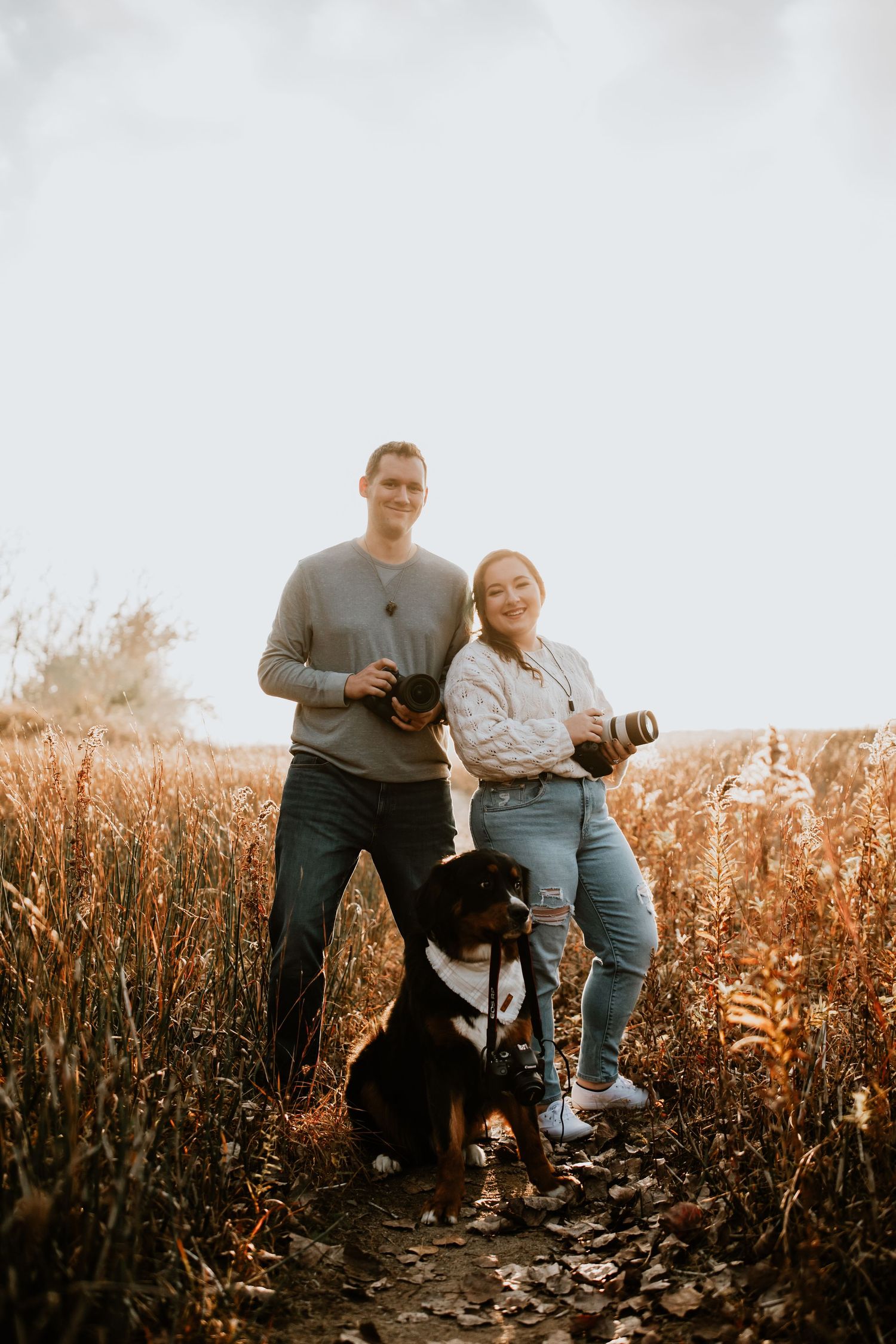 Two photographers with cameras stand in golden field with black dog on rocky trail at sunset.