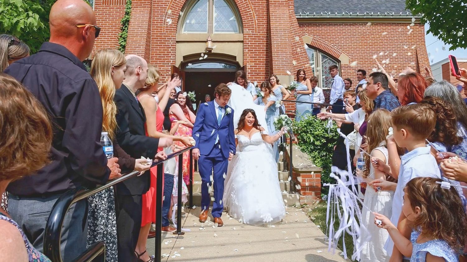 Wedding guests gather outside a red brick church while the newlyweds exit down the steps after their ceremony.